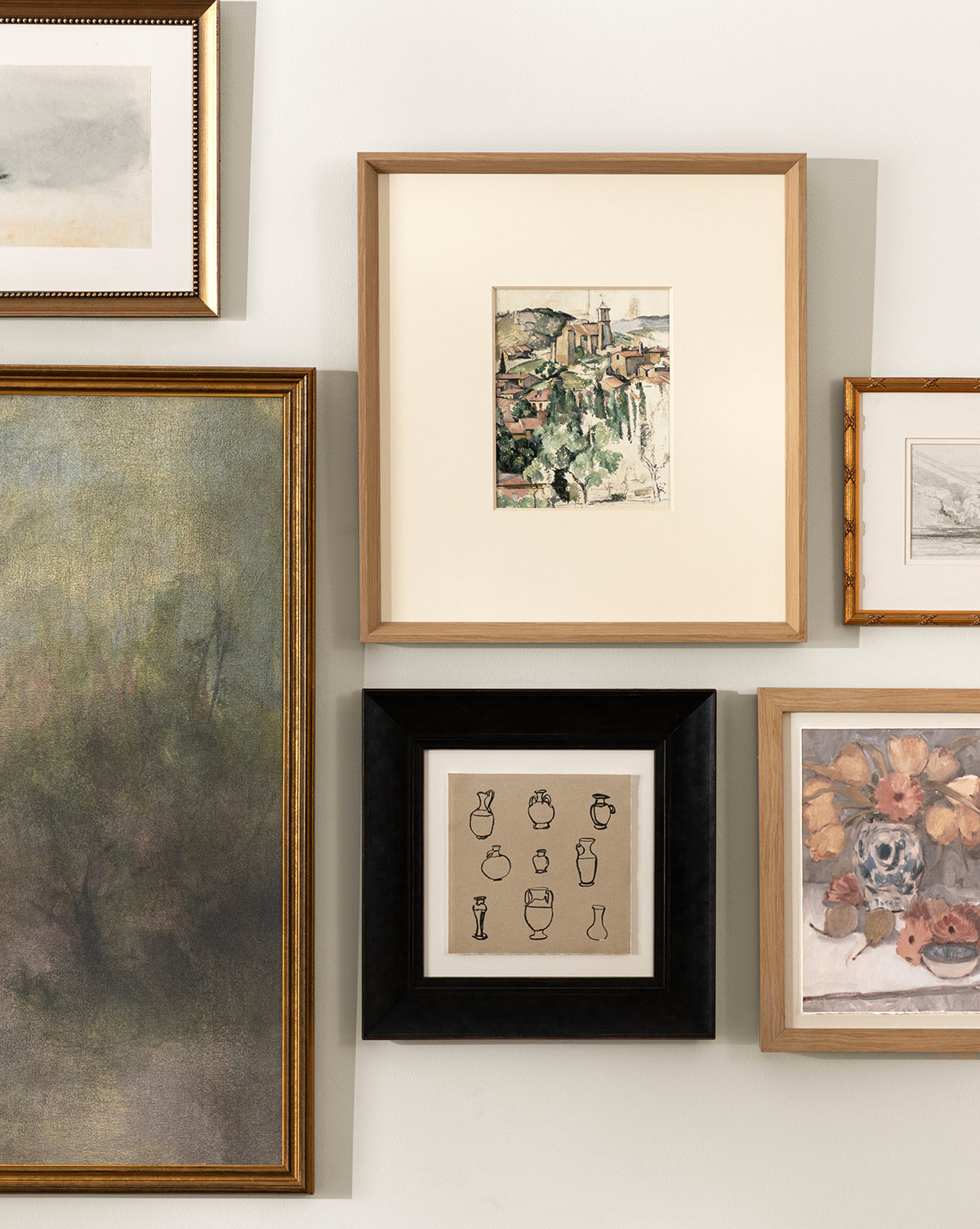 A gallery wall with five Le Village framed artworks, featuring museum-quality giclee prints of landscapes, vases, and scenes arranged as a collage on a light-colored wall in natural woodgrain frames by McGee & Co.