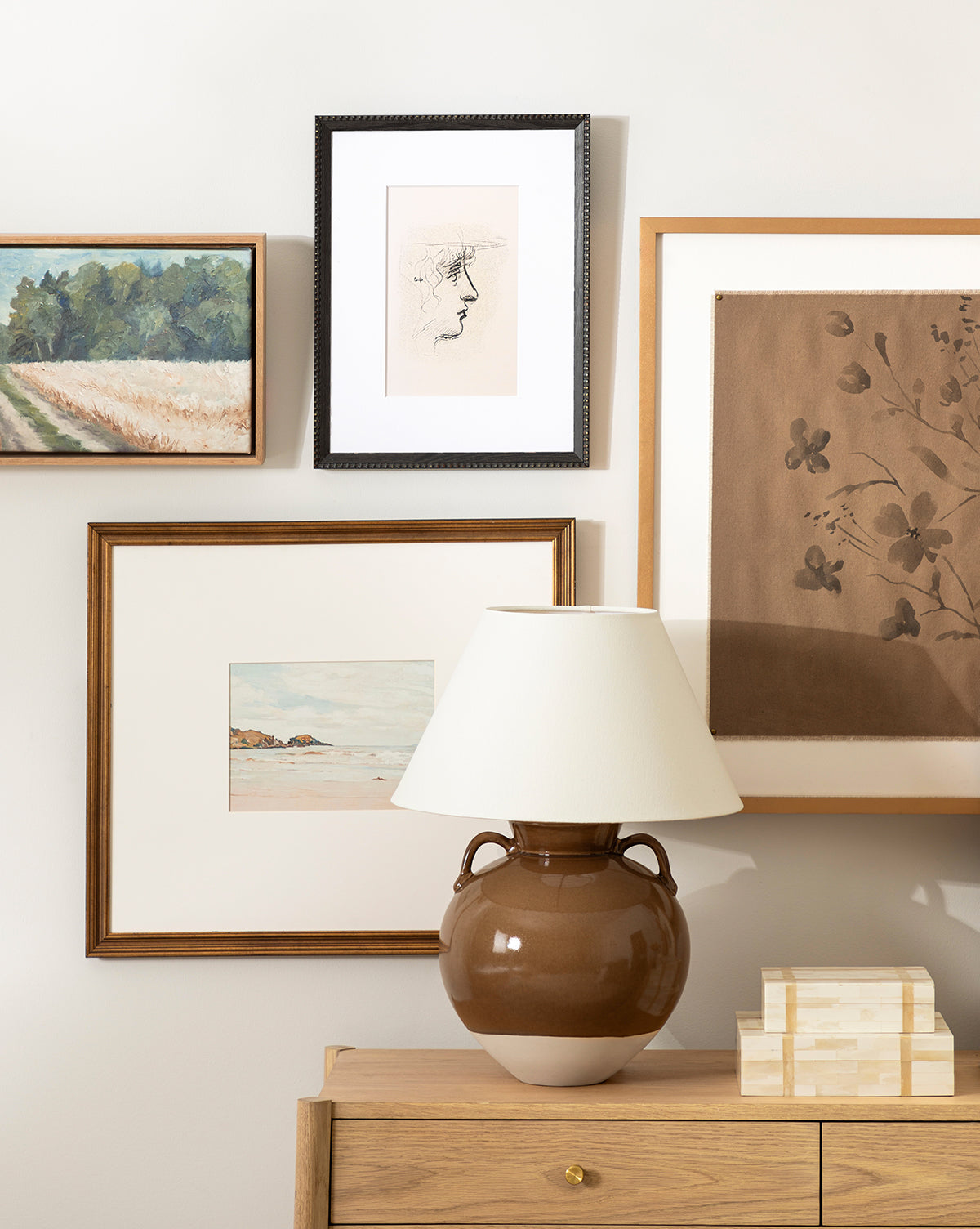 A brown ceramic lamp with a white shade sits on a wooden dresser. Next to it are two stacked Paige Bone Boxes. Framed artwork featuring landscapes and floral prints adorns the light wall behind.