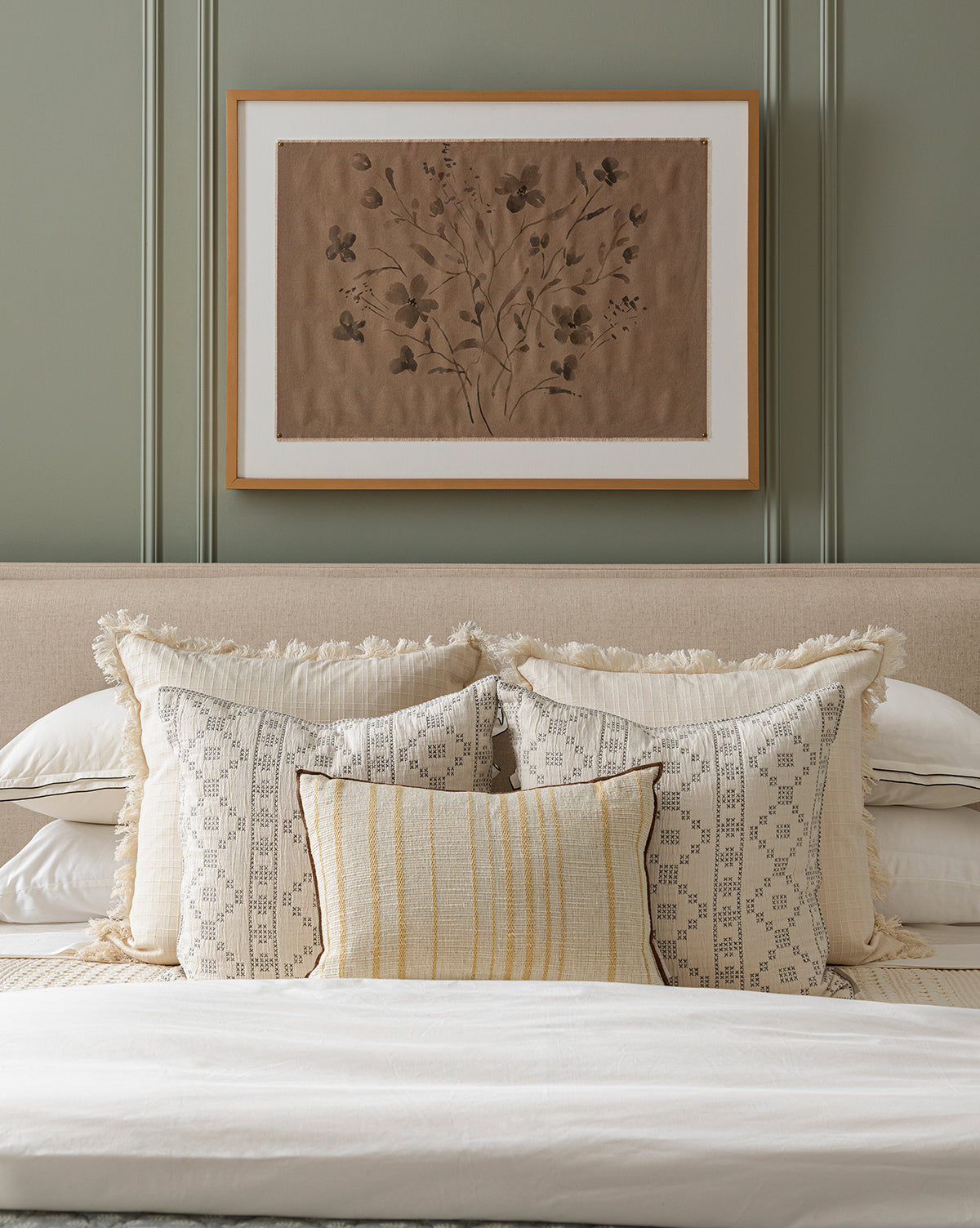 A neatly made bed with cream and beige pillows, a light blanket, and a beige headboard sits under Consider the Wildflowers by Katie Mulder in a maple frame, displayed on a green-paneled wall, by McGee & Co.