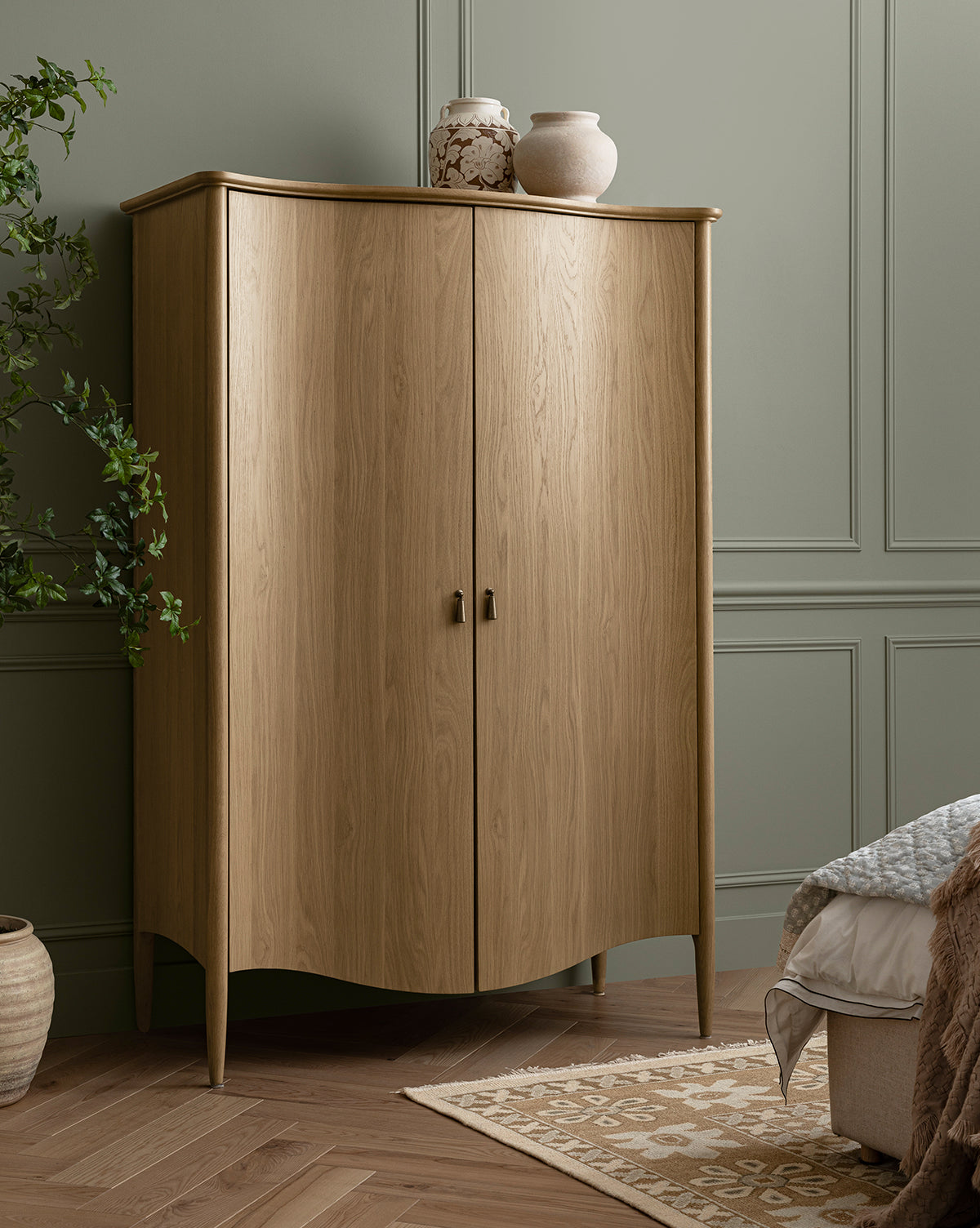 The Moorland Cabinet, a tall light wood piece with adjustable shelves and curved edges, stands against a sage green paneled wall. Decorative vases are displayed on top, with part of a bed and patterned rug visible in the cozy room by McGee & Co.