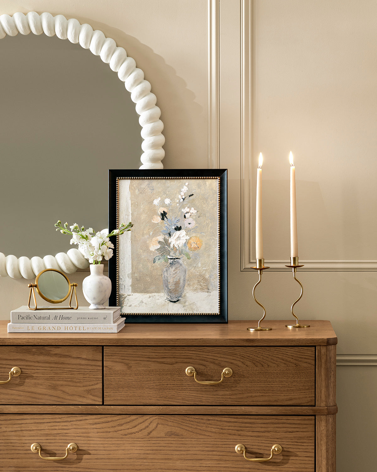 A wooden dresser with gold handles holds two lit brass candlesticks, a framed floral giclee print, stacked books, a small mirror, and the Spring Fresh Cuts vase. A round beaded mirror hangs on the beige wall above.