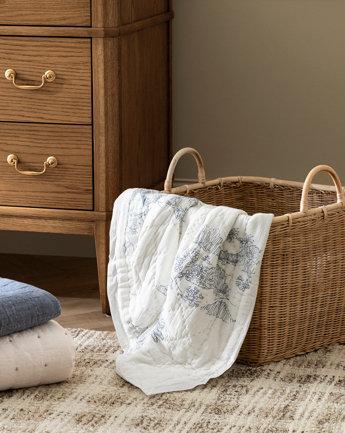 A wicker basket with a Solano Block Print Kids Quilt by Indaba draped over the side sits on a textured rug next to a wooden dresser with brass handles. Folded cotton bedding and kids quilts are stacked neatly on the floor nearby. McGee & Co.