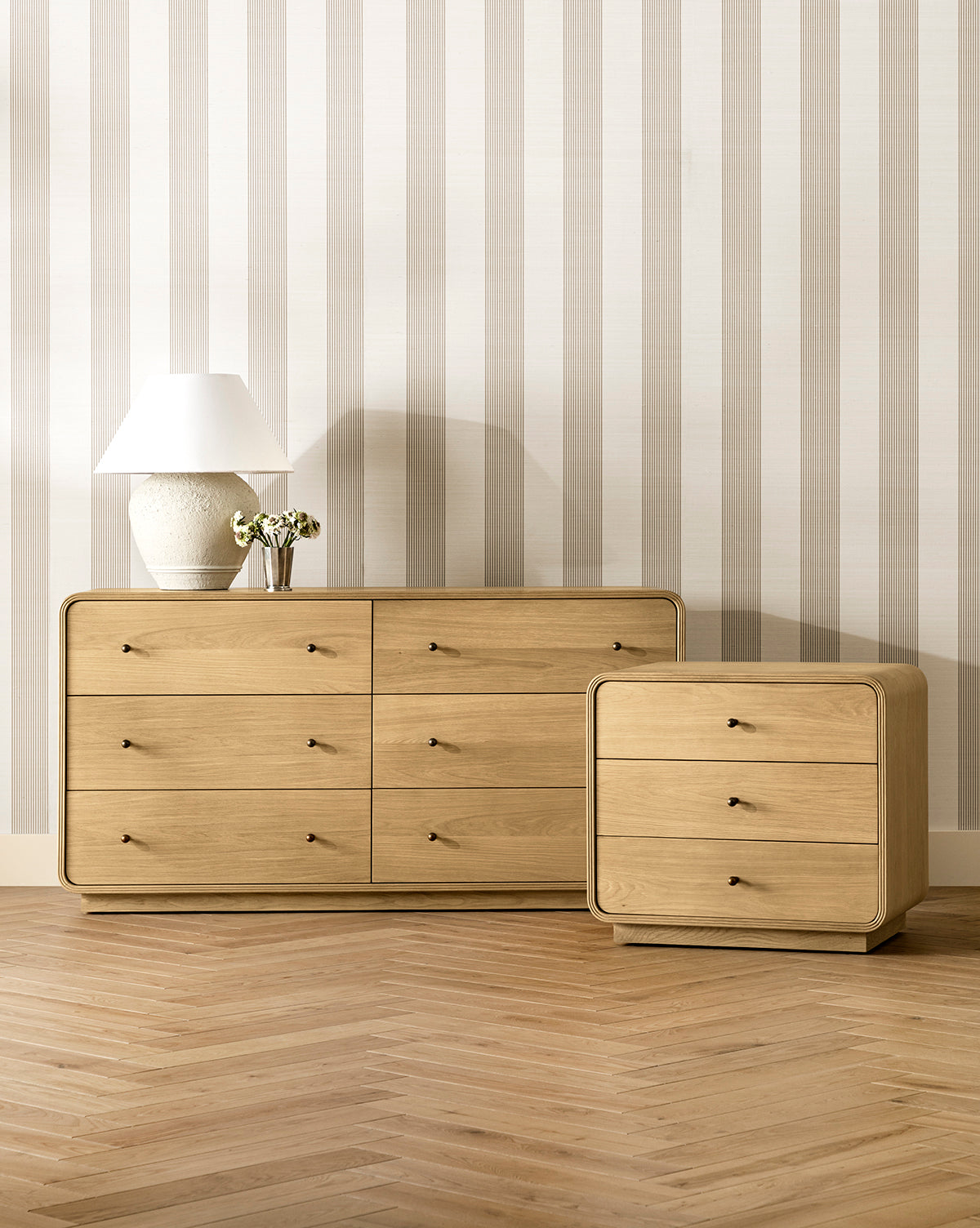 A modern room featuring light wood floors, Leah Striped Sisal Wallpaper by York Wallcoverings, a six-drawer dresser, a matching three-drawer nightstand, a white lamp, and a small vase with flowers by McGee & Co.