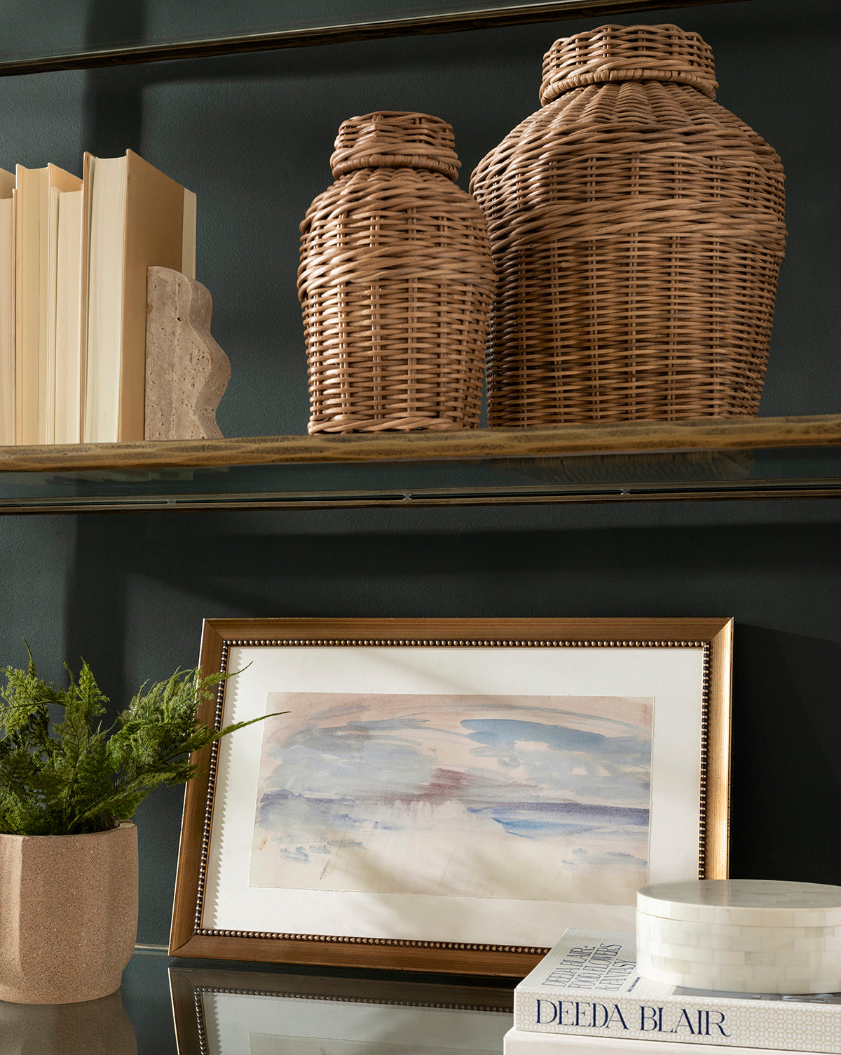 A bookshelf features two woven baskets, books, a potted plant, the calming Sky Studies 3 abstract watercolor giclee print in a frame, and stacked books with a round white box on the lower shelf.