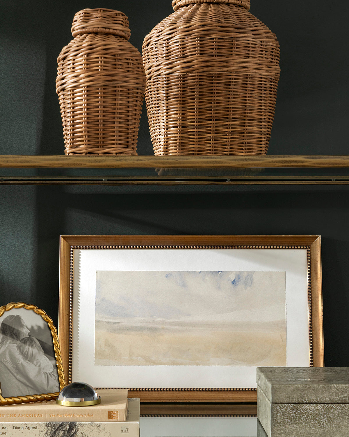 Two woven wicker jars sit on a glass shelf above the calming giclee print Sky Studies 5. Below, books, a small photo in an antique silver wood frame, and a gray box are neatly arranged on a dark background.