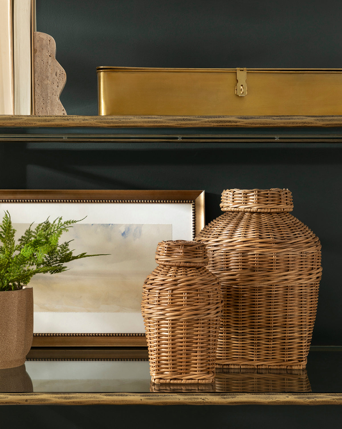 Two Ahnamia Wicker Jars rest on a glass shelf with a framed abstract painting, a small potted plant, and a gold metal box, forming a cozy vignette against the dark background.