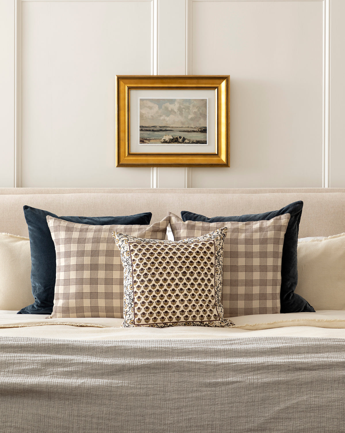 A neatly made bed with beige, plaid, and patterned pillows—including the Bellevue Velvet Pillow Cover—rests against a paneled wall, featuring a gold-framed landscape painting centered above the headboard, by McGee & Co.