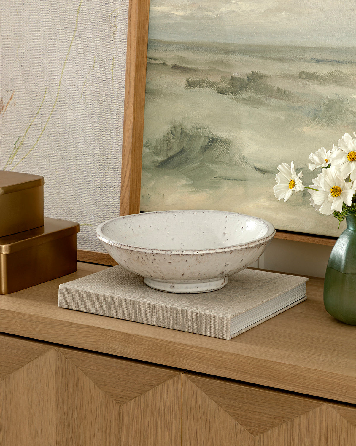 The Kimpton Ceramic Bowl with a white speckled finish sits on a closed book on a wooden cabinet, alongside gold boxes, a green vase with white flowers, and a framed landscape painting in the background, by McGee & Co.