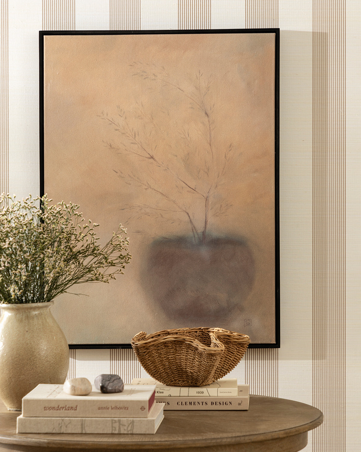 A tabletop styled with a ceramic vase, stones on stacked books, and the Goulding Woven Bowl. On the striped wall behind, a soft painting of a vase with dry grasses completes this serene entryway decor by McGee & Co.
