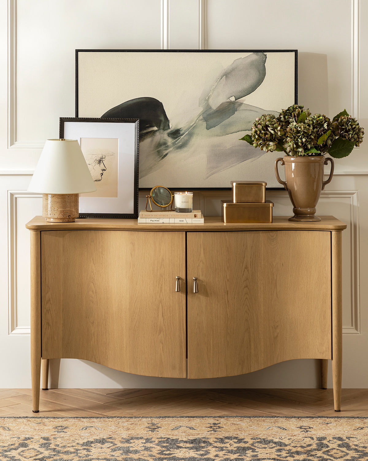 A wooden sideboard stands against a paneled wall, topped with a lamp, gold boxes, candle, clock, vase of green hydrangeas, and framed Head Study artwork featuring expressive linework. A patterned rug covers the floor by McGee & Co.
