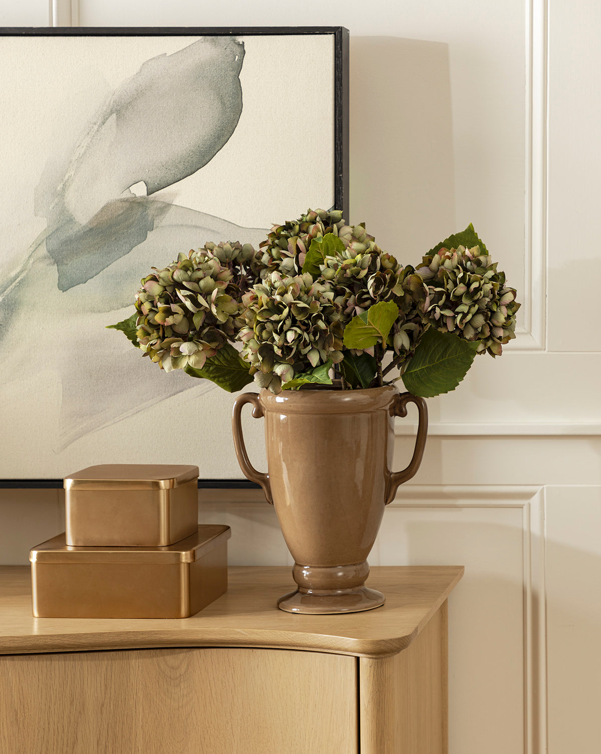 A brown ceramic vase with two handles holds green hydrangeas on a light wood table beside the Oswald Metal Box, which has a brass finish, and another gold box, set against abstract framed artwork in the background.