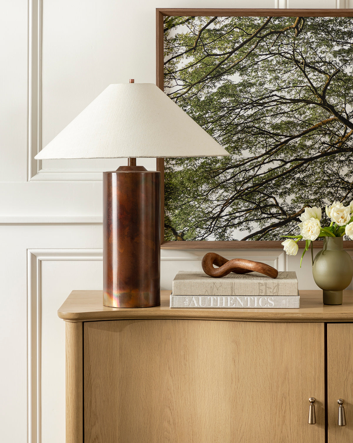 A Seaton Table Lamp with a metal base sits on a wooden sideboard alongside a framed tree photo, stacked books, a brown sculpture, and white flowers in a vase, all set against a white paneled wall for an industrial look, by McGee & Co.