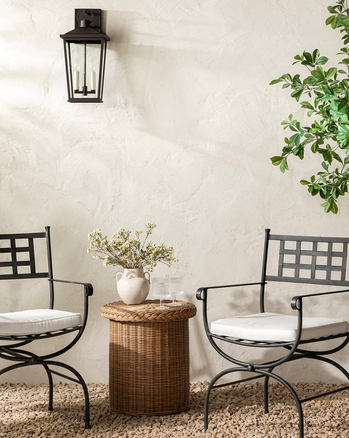 Two black Addie Outdoor Chairs with performance fabric cushions face a round wicker table topped with flowers, water glasses, and a book, set against a textured white wall featuring a lantern and lush leafy plant by McGee & Co.