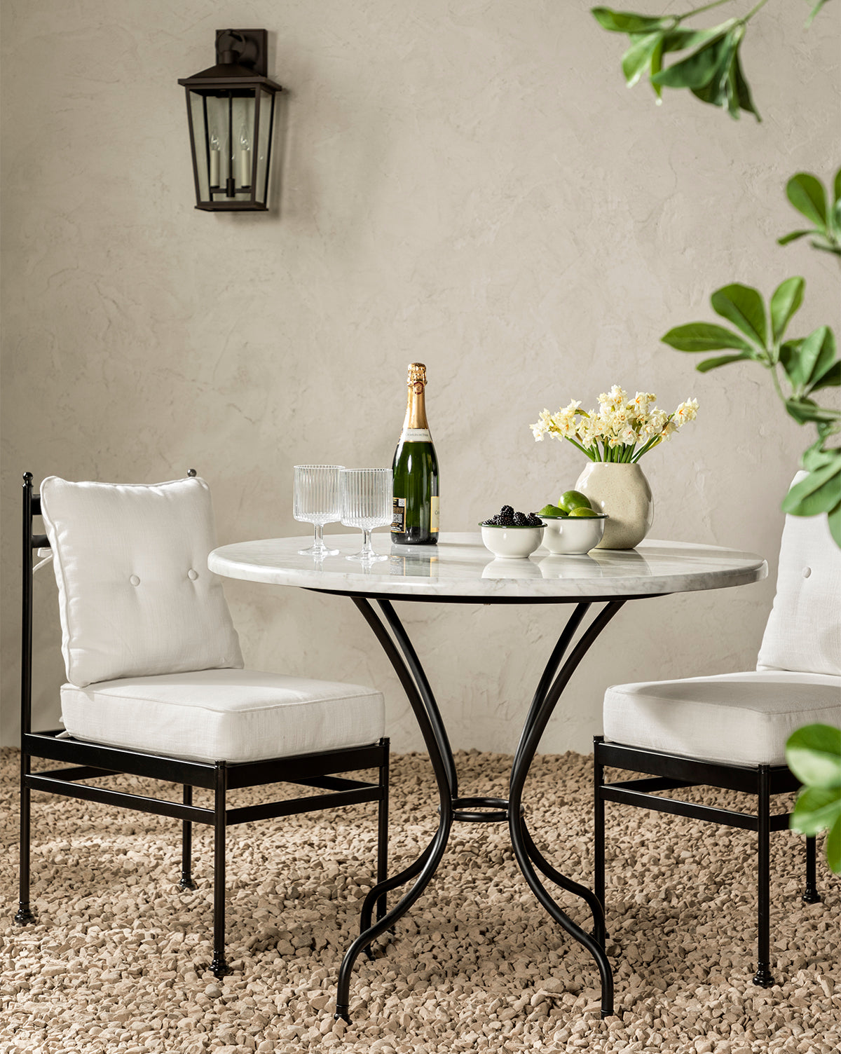A round marble table with two Pomeroy Outdoor Dining Chairs in Pasadena White performance fabric sits on a gravel floor, topped with champagne, glasses, a flower vase, black olives, green apples, and a lantern hanging nearby by McGee & Co.