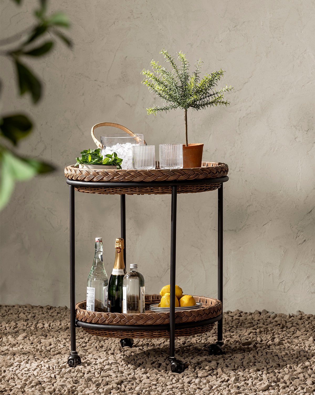 A wicker and metal bar cart on pebbled ground holds bottles, glasses, lemons, a terracotta pot with a Potted Faux Rosemary Topiary, and a basket of ice against a textured beige wall with leafy branches in the foreground.