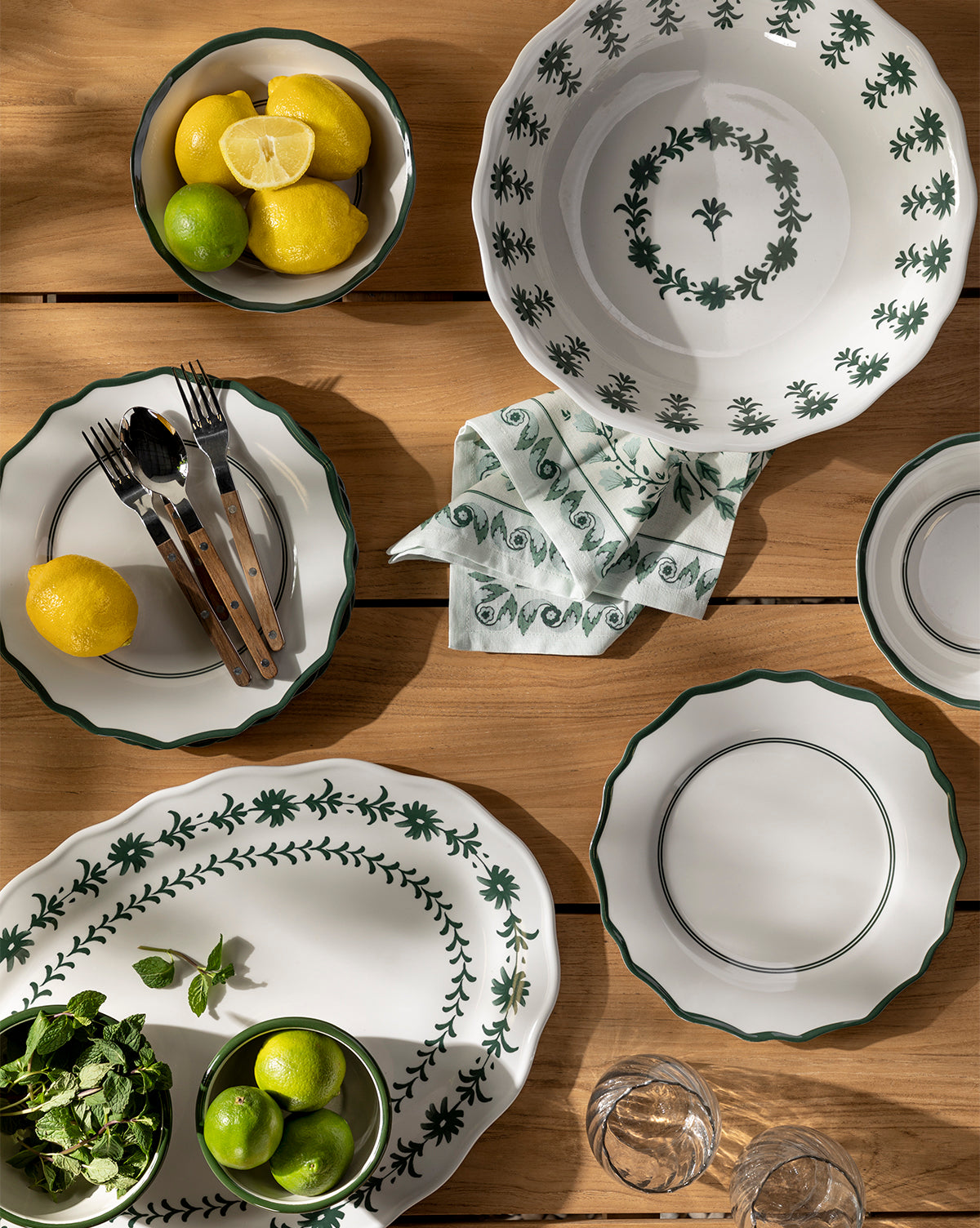 A wooden tabletop displays white dishes with a green floral motif, bowls of lemons and limes, herb sprigs, cutlery, napkin, glasses, and the Alba Melamine Serving Bowl with a scalloped edge, by McGee & Co.