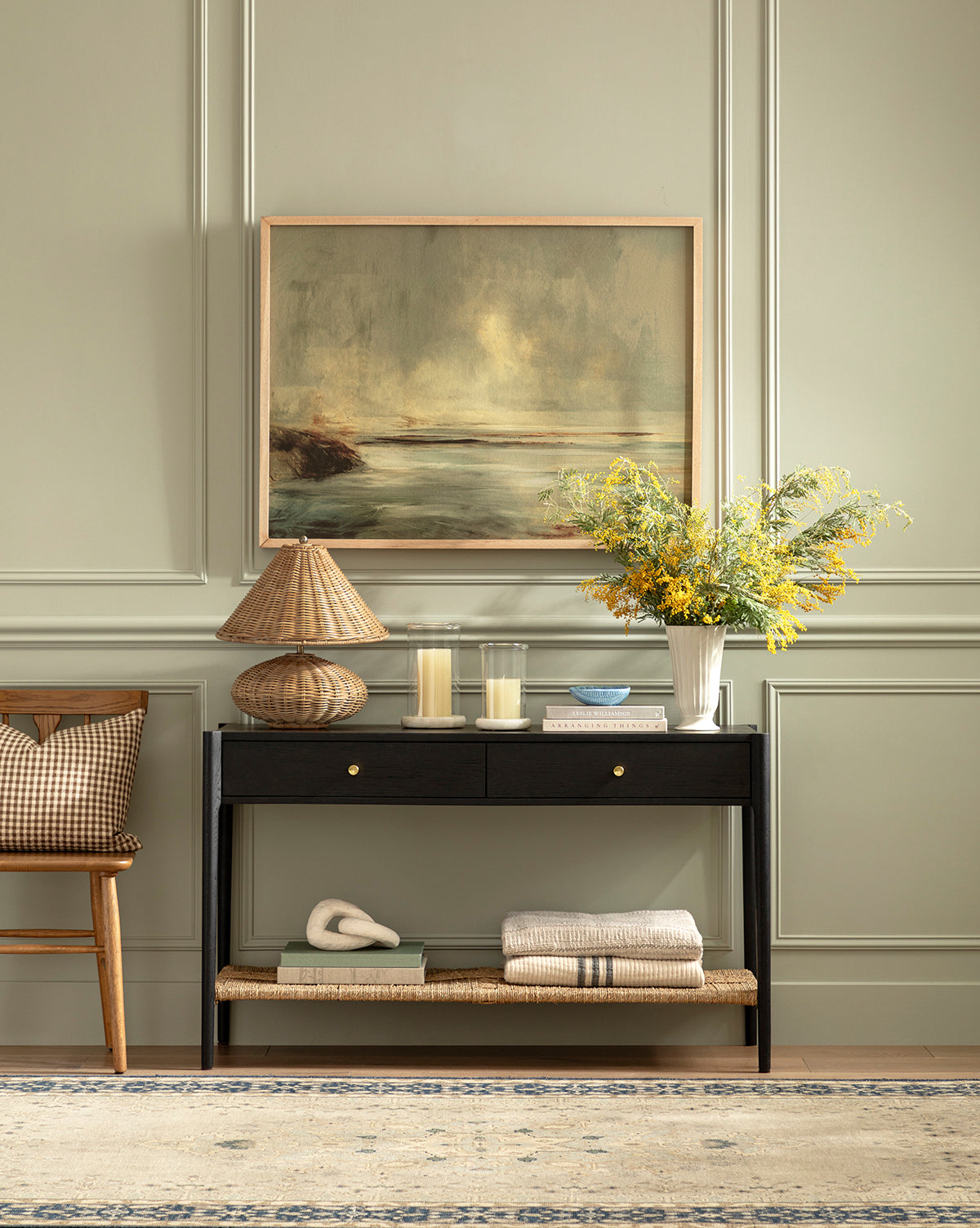 The Dana 2-Drawer Console, topped with a lamp, candles, and a yellow flower vase, stands against a pale green paneled wall beneath a framed landscape—creating an elegant entryway beside a chair and folded blankets by McGee & Co.