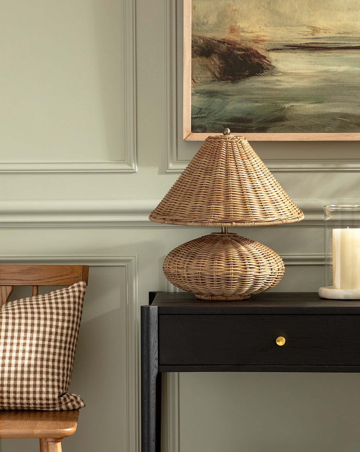 The Palmetto Table Lamp by Paul-Yu Industrial Corporation sits on a black console table beside a large candle in a glass holder. A wooden chair with a brown checkered cushion is partially visible, beneath an abstract landscape painting on a green paneled wall. McGee & Co.