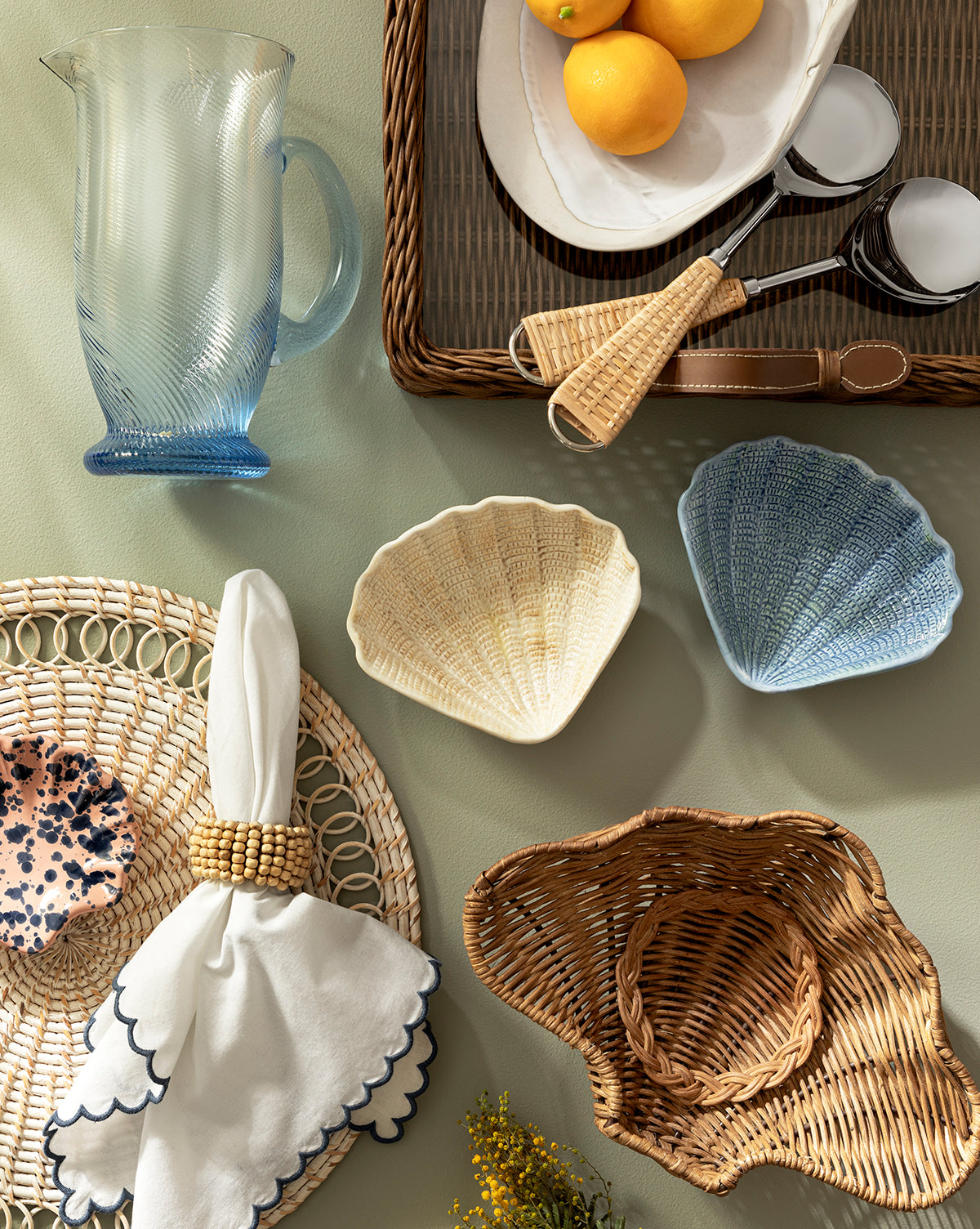 A flat lay of summer tabletop essentials featuring the Spiral Jug, a rattan tray with lemons and ice tongs, shell-shaped bowls, woven placemats, a napkin with a beaded ring, and a woven sunhat on a light green surface by McGee & Co.