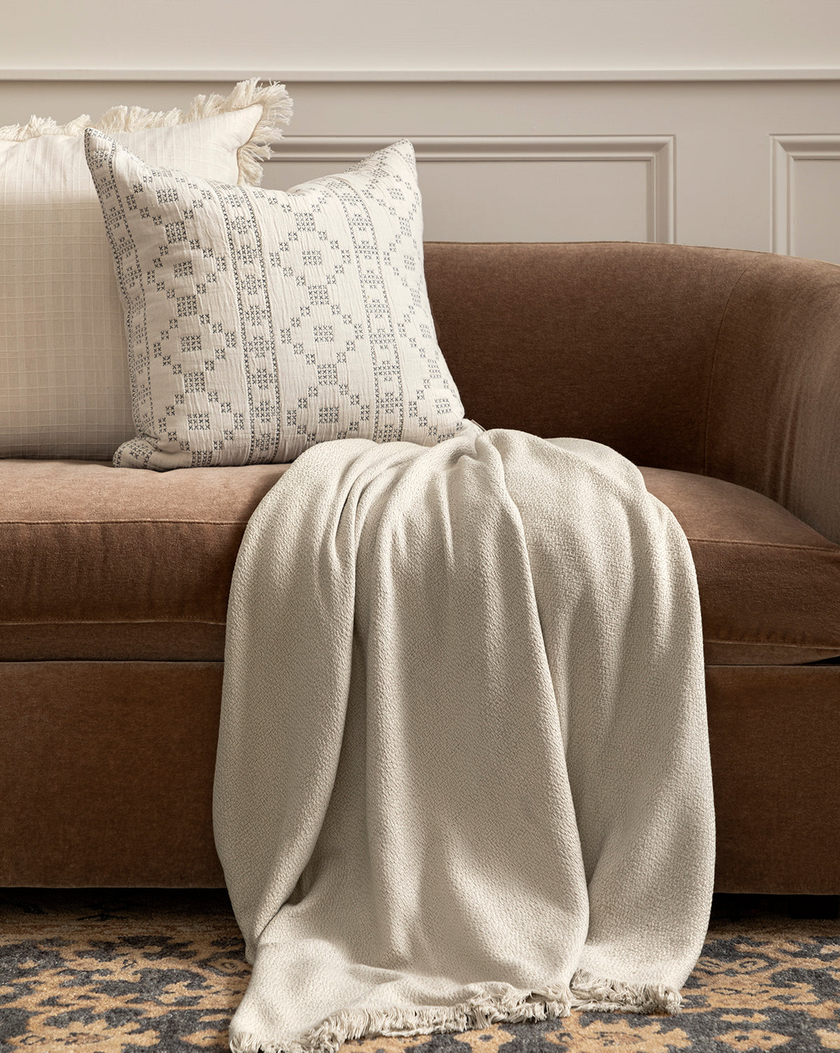 A brown velvet sofa features the Ariella Throw by J Call Designs LLC dba Sew Miami LLC draped over the seat, with a patterned pillow at the armrest. Behind, a white wall with decorative molding completes the neutral palette, complemented by an accent from McGee & Co.