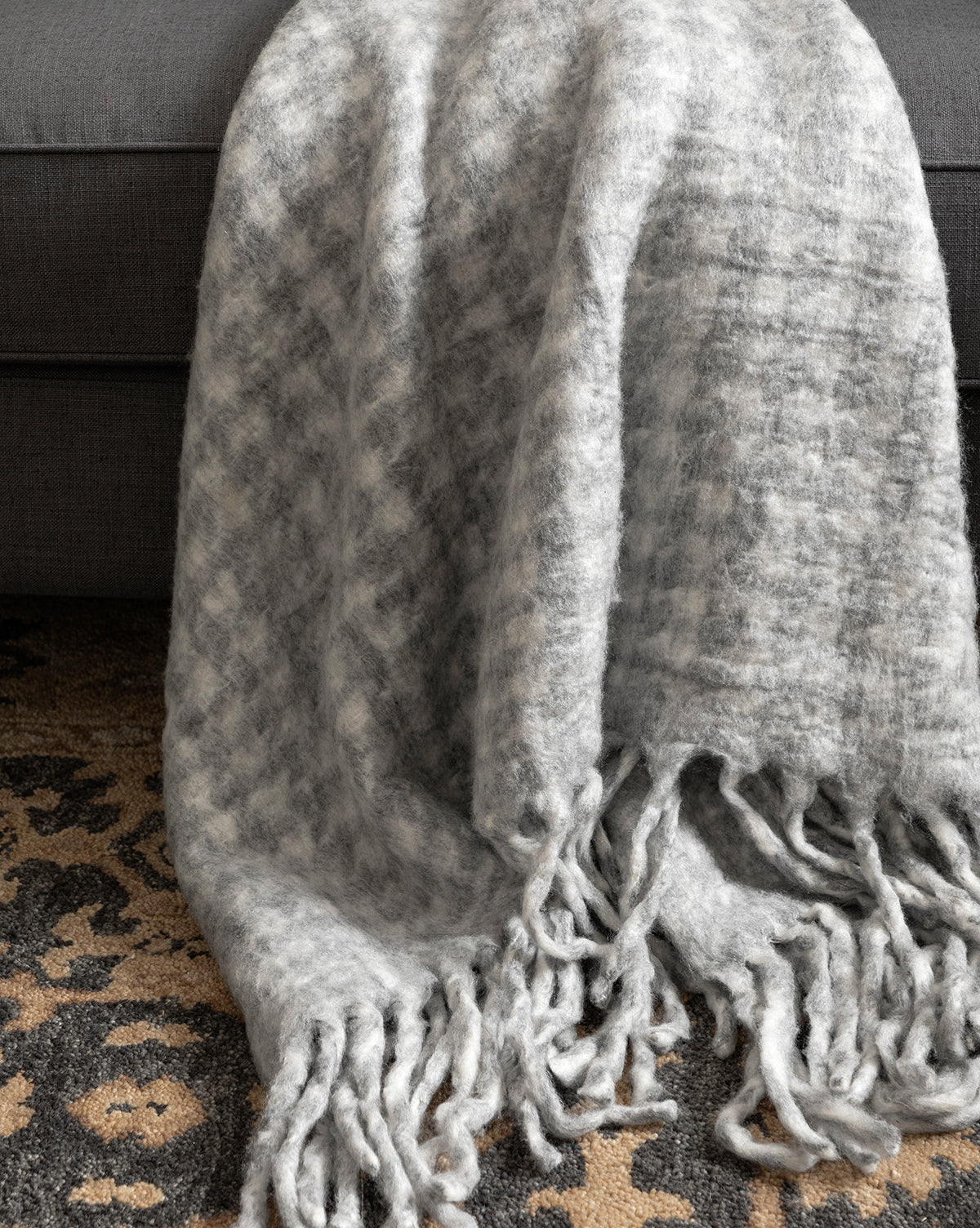 The Indaba Kirsley Houndstooth Throw, in soft gray and white wool blend with fringe, elegantly drapes over a dark gray sofa on a black and tan patterned carpet—an ideal touch for luxury home decor. McGee & Co.