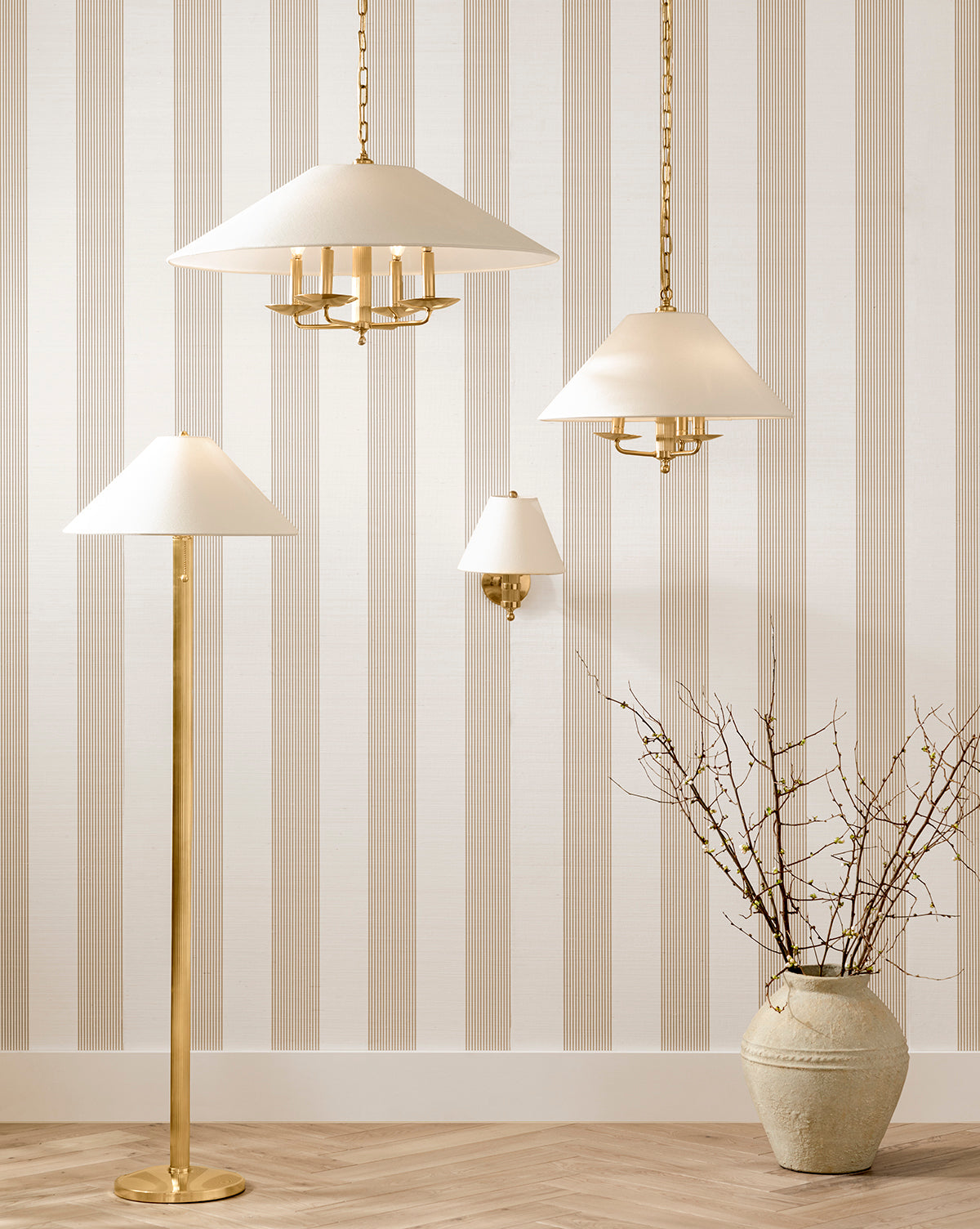 Four elegant gold and white lamps with linen shades—three hanging, the Townes Floor Lamp in reeded brass, and a wall sconce—are set against beige-and-white striped wallpaper beside a large vase with branches on a herringbone wood floor, by McGee & Co.