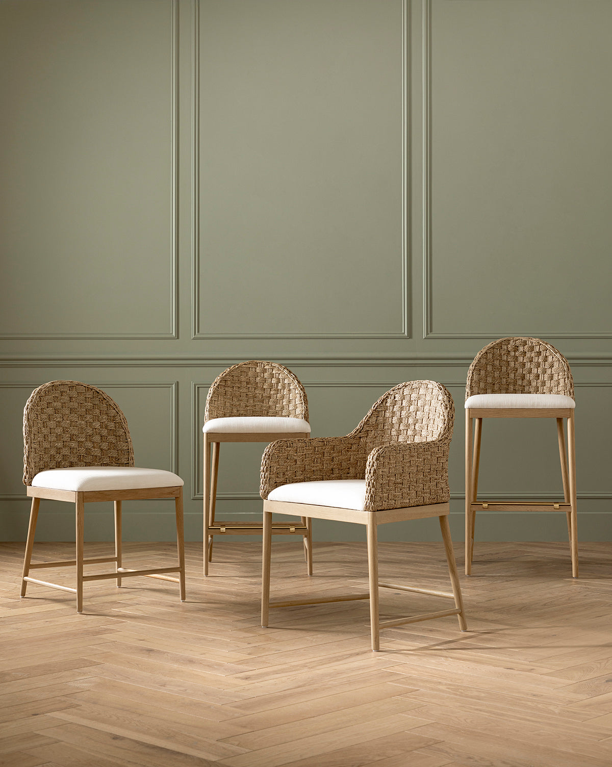Four Molly Arm Chairs with woven rattan backs and white cushions are set on a herringbone wood floor, placed against a sage green paneled wall by McGee & Co.