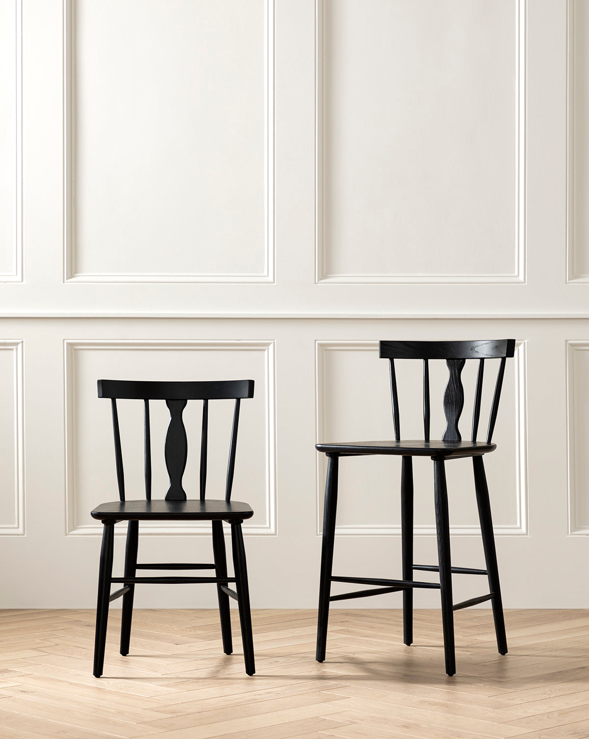Two black wooden chairs, including a Whittaker Counter Stool by YANG CHENG WOODEN INDUSTRIES INTERNATIONAL (VIETNAM) CO LTD, with a spindle back design, sit side by side on a herringbone wood floor against an off-white paneled wall. McGee & Co.