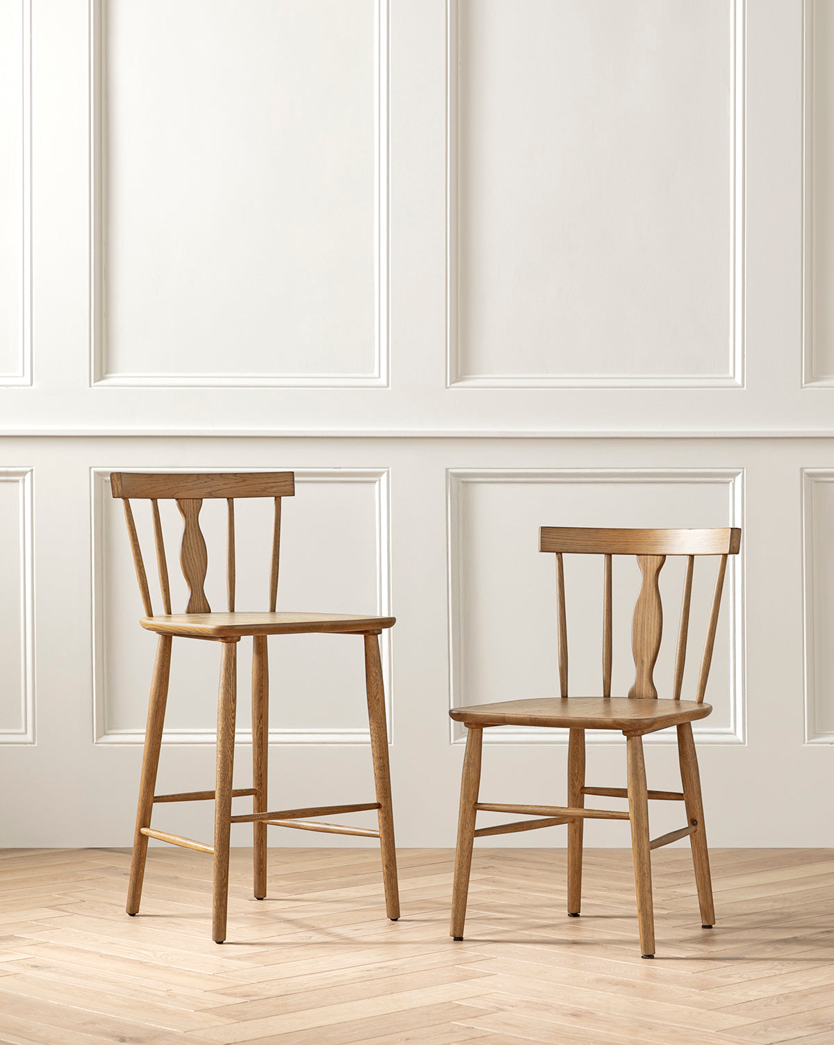 A Whittaker Counter Stool and a matching chair by YANG CHENG WOODEN INDUSTRIES INTERNATIONAL (VIETNAM) CO LTD stand before a white paneled wall on a light herringbone wood floor, both with simple spindle backs and solid oak, light brown finishes. McGee & Co.
