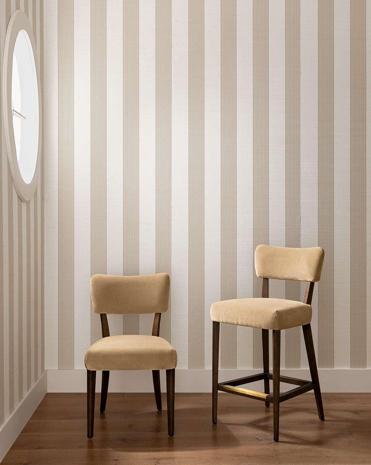 Two Etienne Dining Chairs (Ready to Ship) in beige—one shorter, one taller—are placed on a wooden floor against a vertically striped beige and white wall, with a round window at left highlighting this vintage-inspired dining setup by McGee & Co.