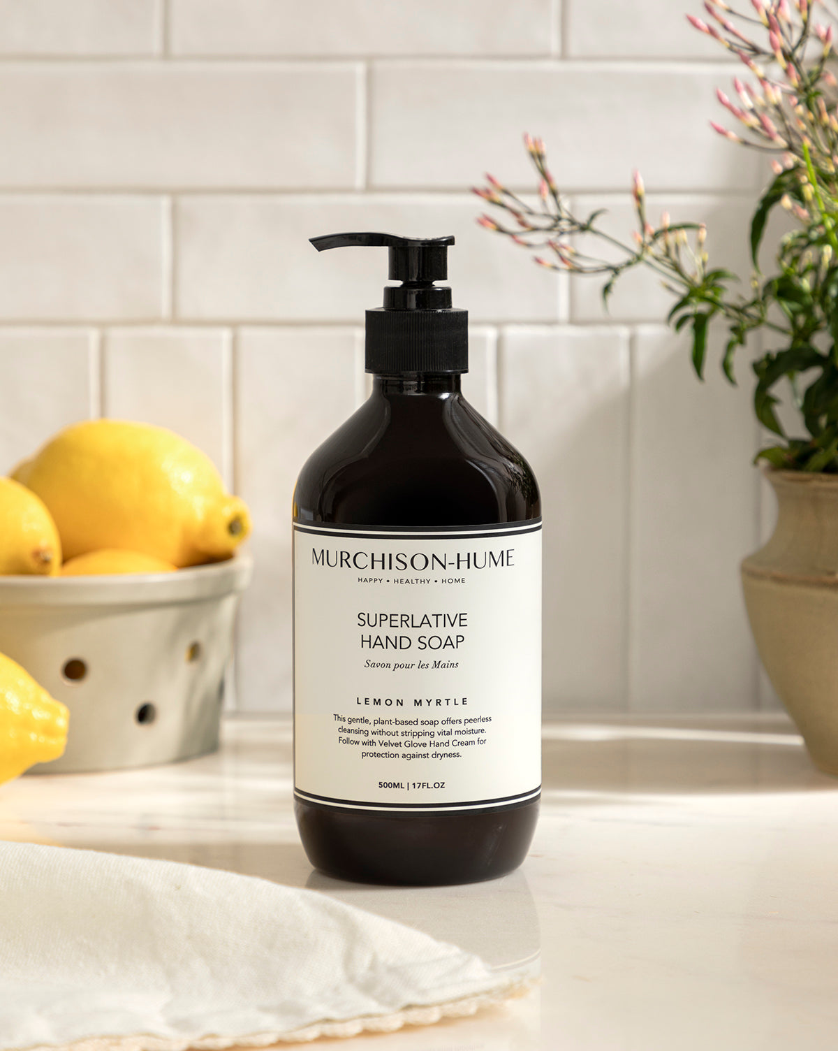 A bottle of Murchison-Hume Hand Soap with shea butter in lemon myrtle scent rests on a kitchen counter, surrounded by lemons and leafy branches, set against a white tile backsplash.