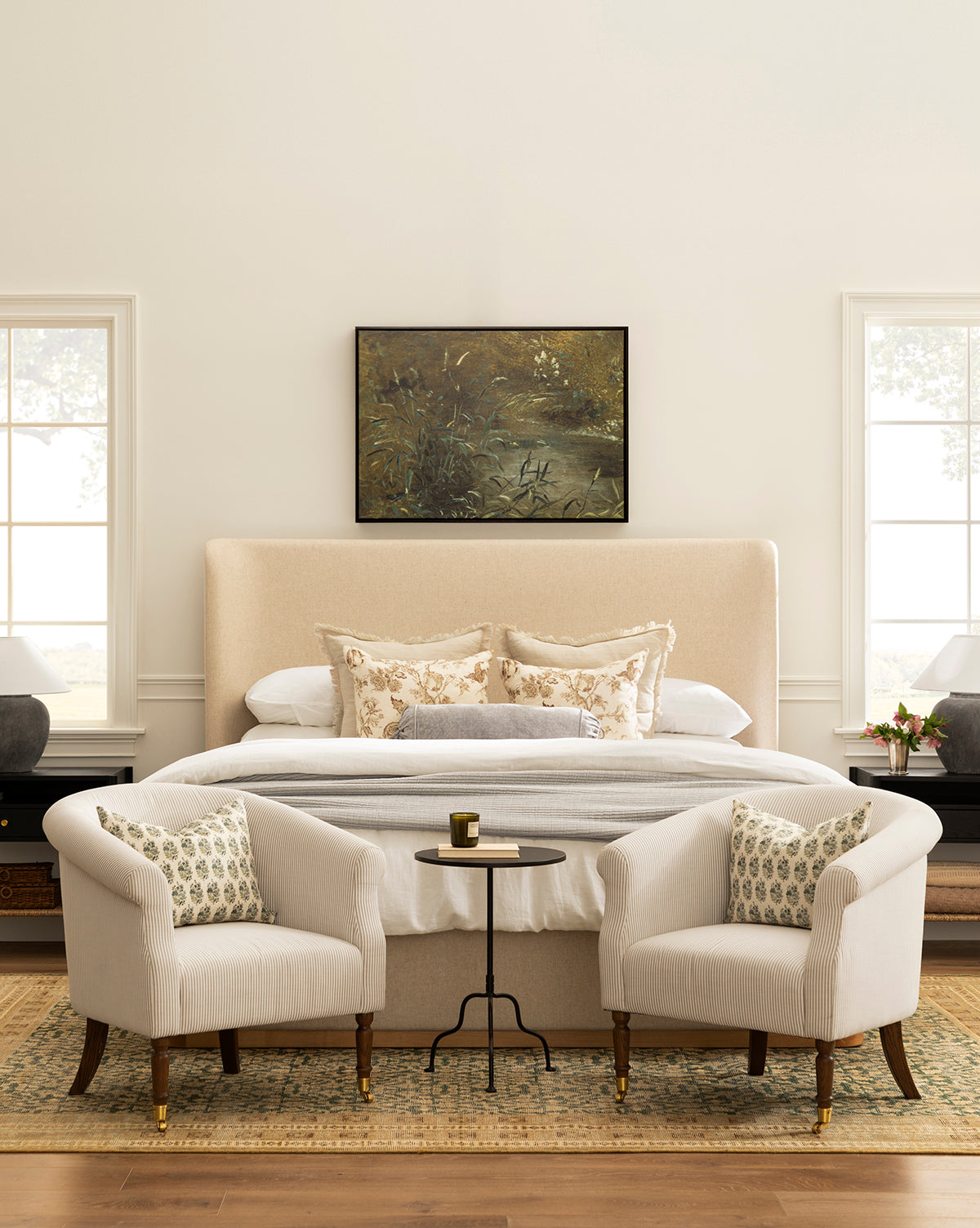 Elegant bedroom with a beige upholstered bed, floral pillows, two white armchairs with patterned cushions, and a small round table. Above the bed hangs Bank Grasses gallery-wrapped giclee on canvas. Large windows fill the room with natural light, by McGee & Co.