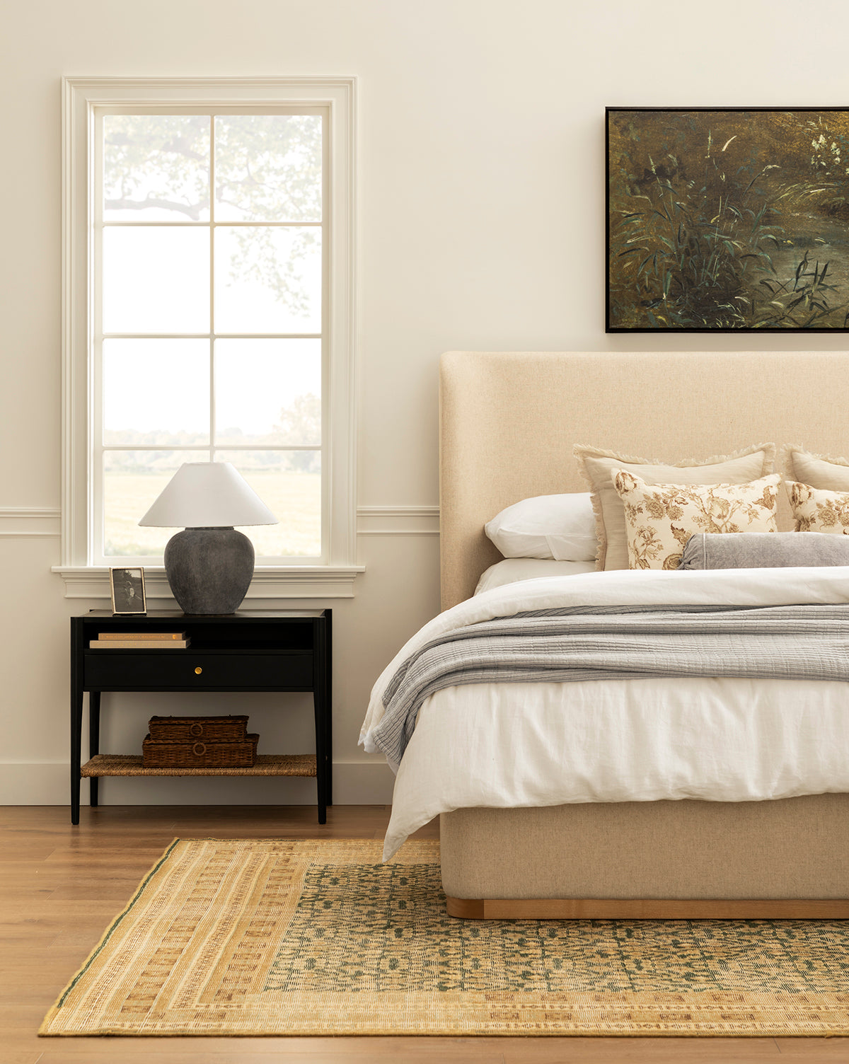 A cozy bedroom with a beige upholstered bed, white and floral pillows, gray lamp on the Dana Nightstand featuring a black finish and woven seagrass shelf, wicker baskets below, window view, yellow-patterned rug, and nature painting by McGee & Co.