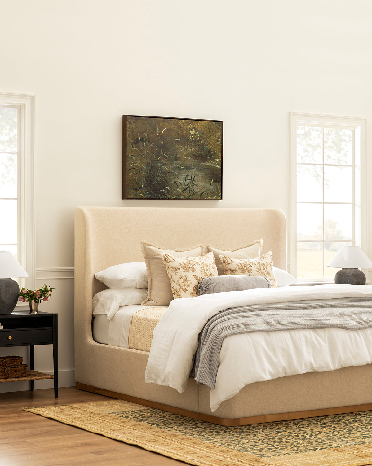 A cozy bedroom with the Denning Upholstered Bed by Makerspalm in beige, layered neutral bedding, floral pillows, two black nightstands with lamps, a yellow woven rug, large windows, and a nature painting above the bed. McGee & Co.