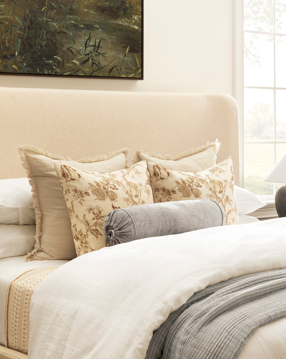 A neatly made bed features cream and beige floral pillows, the Delphine Bolster Pillow in gray stonewashed cotton, light bedding, and a beige upholstered headboard. A large window and vintage-inspired landscape painting appear in the background.