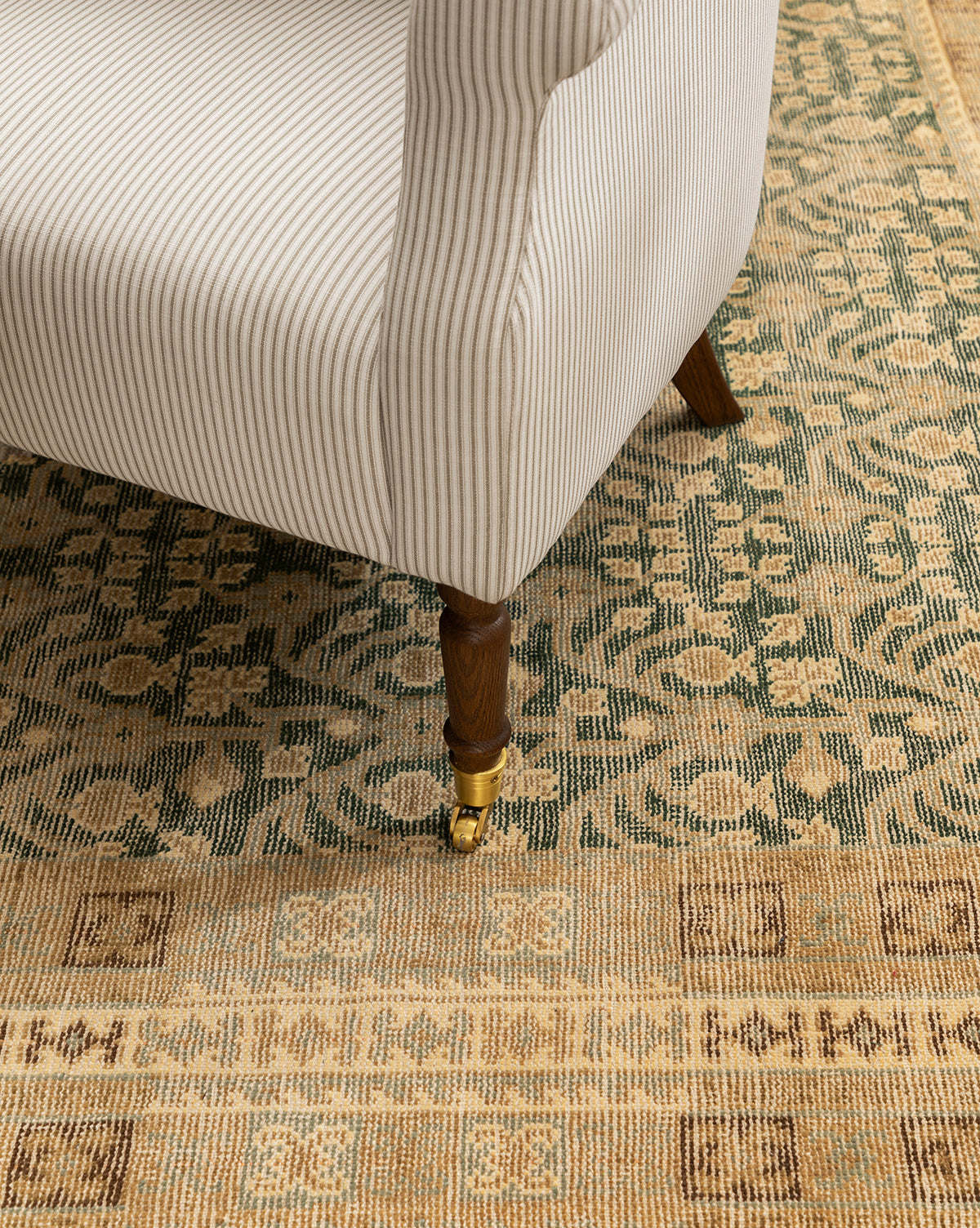 A close-up of the Clemence Lounge Chair’s leg with beige and white striped upholstery and a brass caster, set on a patterned beige and green area rug, by McGee & Co.