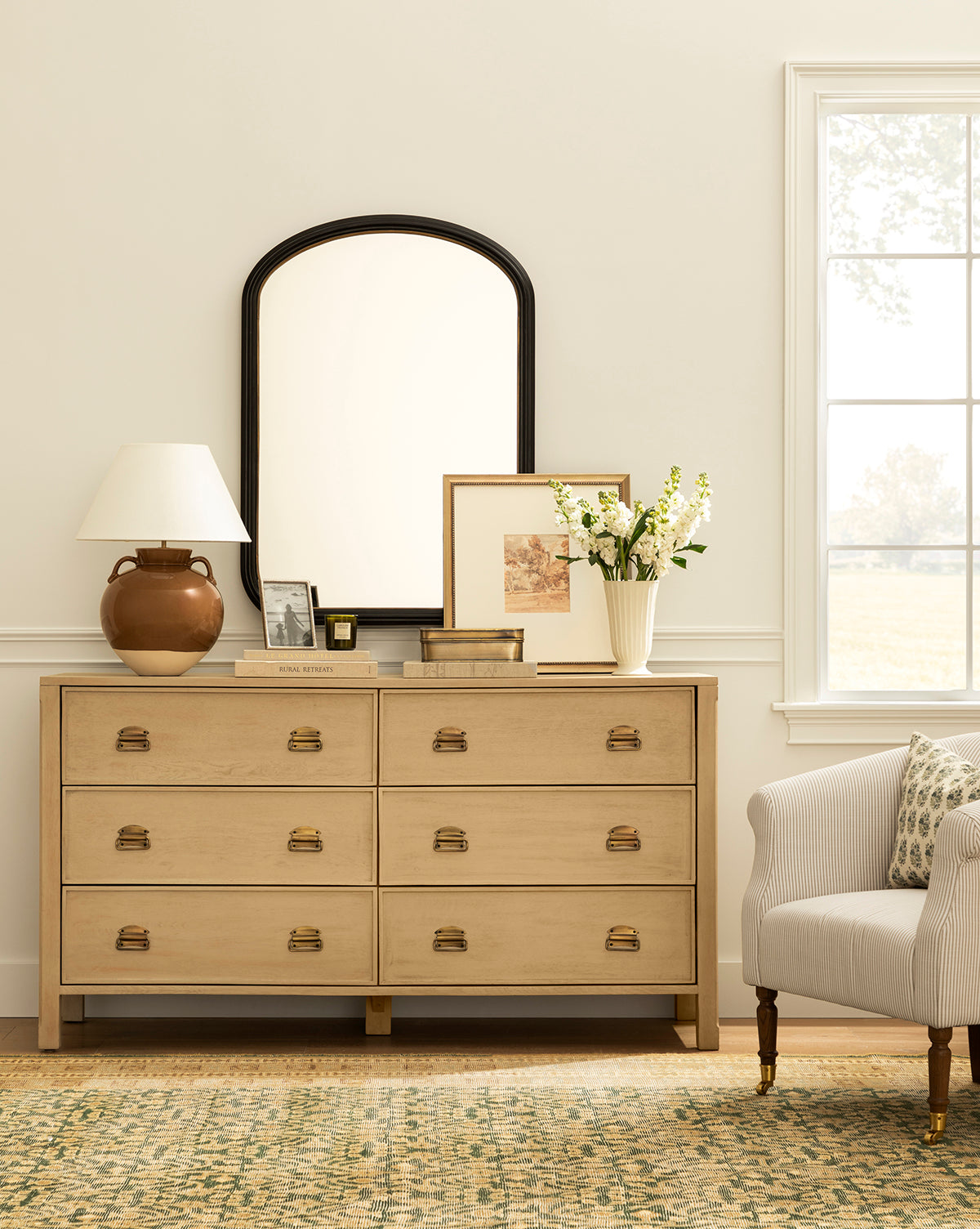 A light wood dresser with gold handles displays a brown lamp, a floral vase, framed art, and books. Above it hangs the Samuel Wall Mirror. To the right are an upholstered armchair and a sunlit window by McGee & Co.