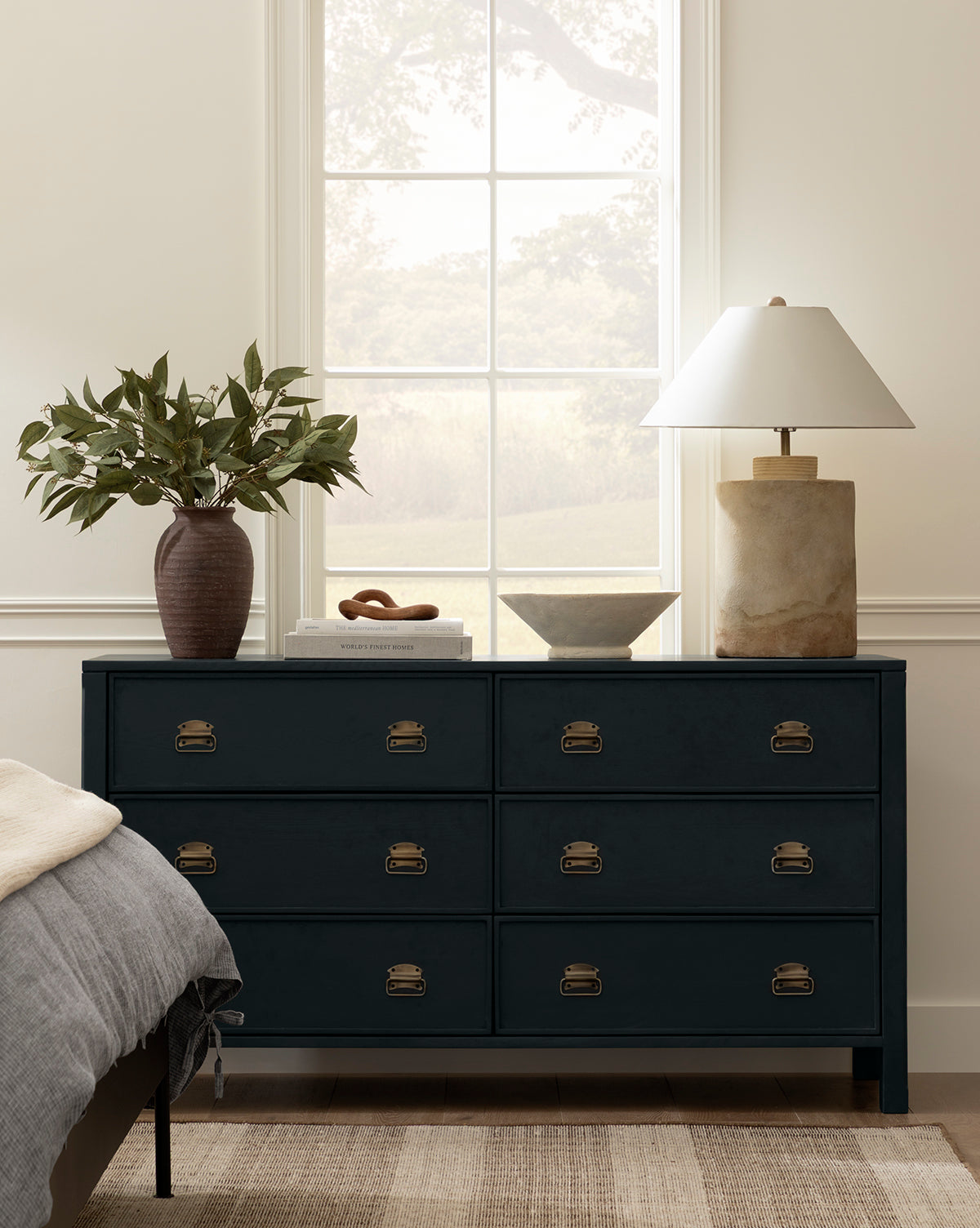 A navy blue dresser with brass handles stands against a cream wall beneath a large window. On top sit green leafy branches in a brown vase, the Matias Bowl in weathered white, and a beige marble lamp with a white shade; part of a bed is visible.