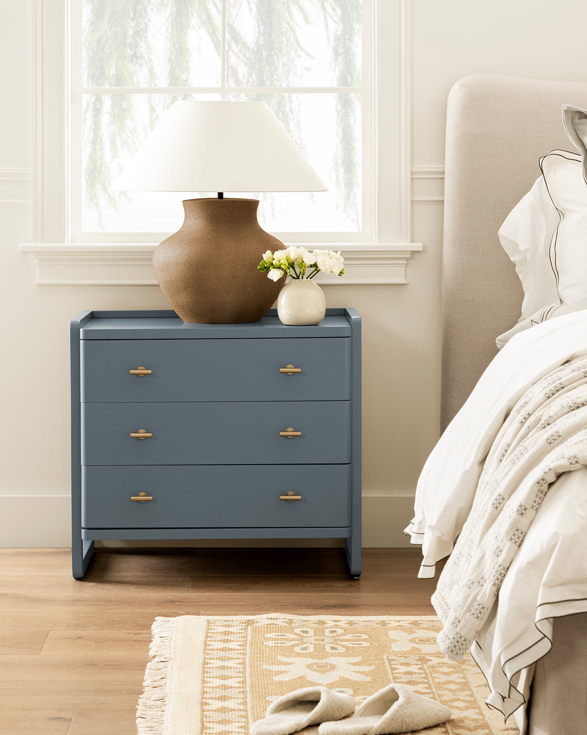 The Butler Nightstand in slate blue with four drawers is placed next to a beige upholstered bed. On top, there’s a large ceramic lamp, small white vase with flowers, and a decorative bowl. A window is also visible nearby by McGee & Co.