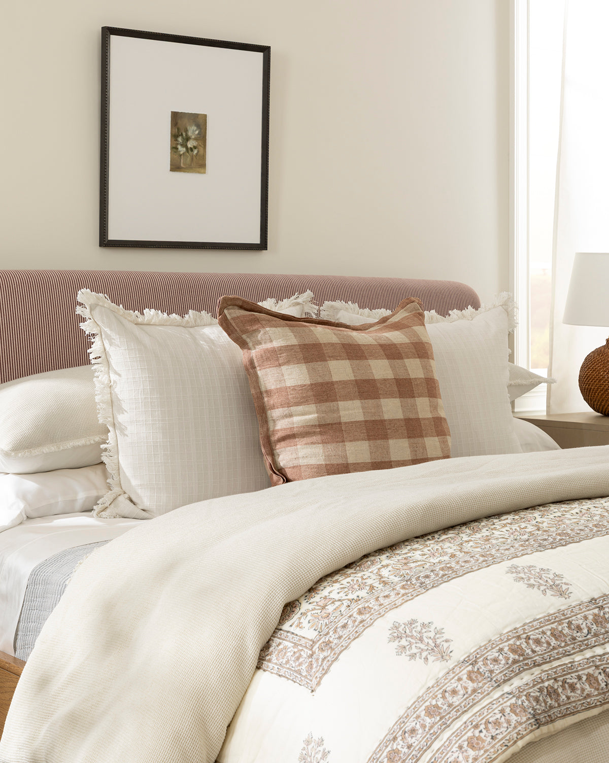 A bed with white ruffled pillows, a brown plaid pillow, and a cream patterned comforter sits beneath Flower Vase Still Life by Katie Mulder in a wood frame, displayed on a light wall as sunlight streams in, by McGee & Co.