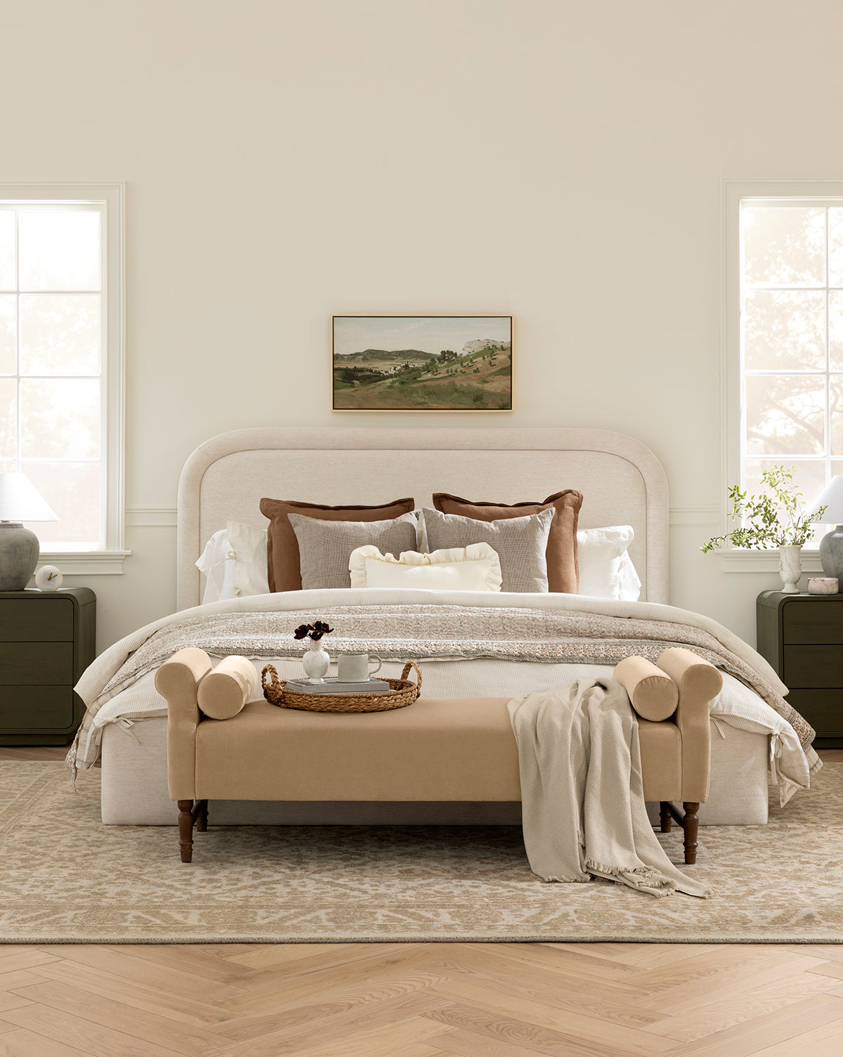A cozy bedroom features a cream upholstered bed with striped linen bedding, brown and white accent pillows, green nightstands, table lamps, a landscape painting above the bed, and the Asher Bench in beige at the foot, by McGee & Co. Sunlight pours in from two windows.