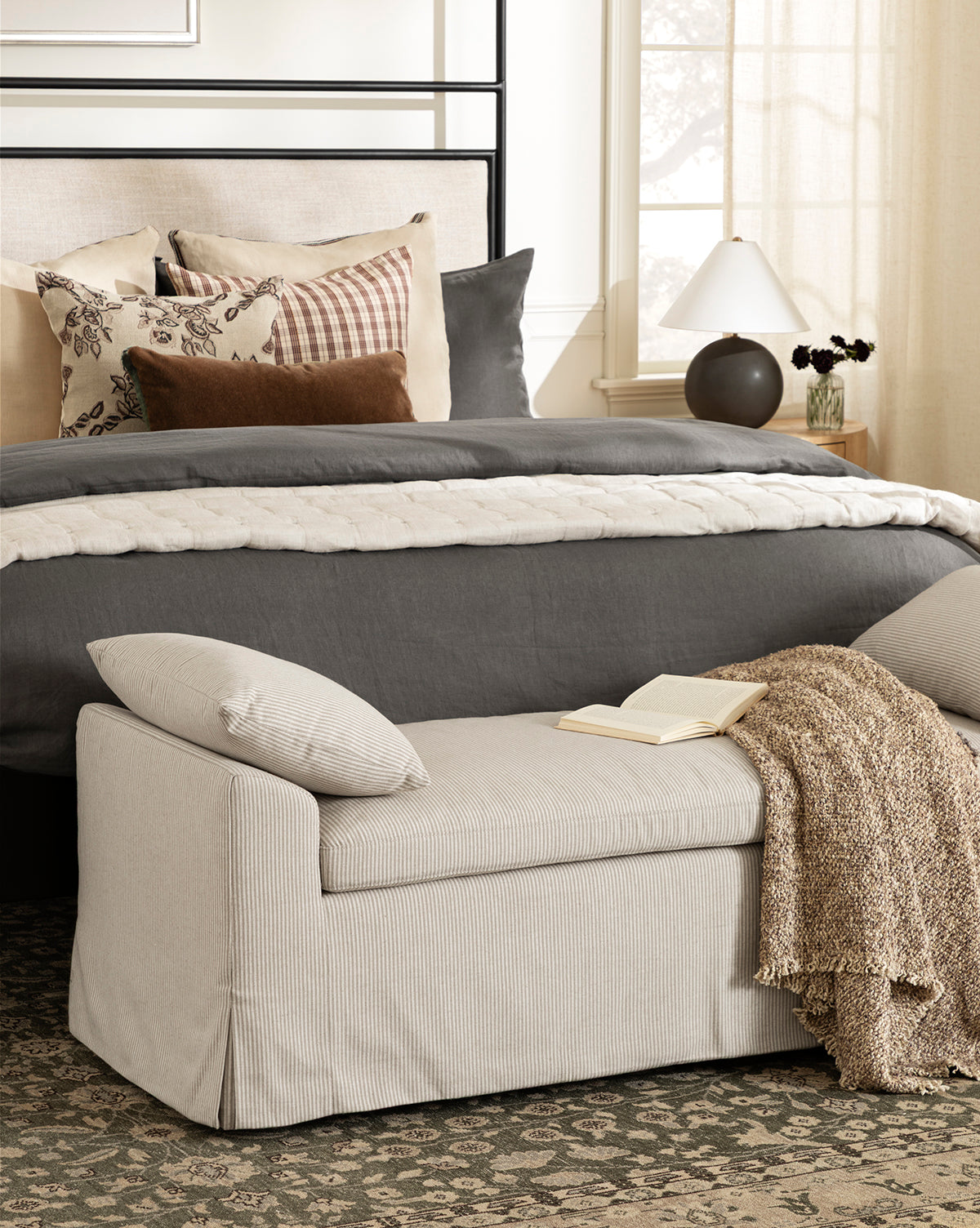 A cozy bedroom with a neatly made bed in gray and beige, assorted pillows, a patterned rug, and a Ready to Ship Scaletta Bench with linen upholstery—topped with a pillow, throw, open book—plus a nightstand with lamp and vase by McGee & Co.