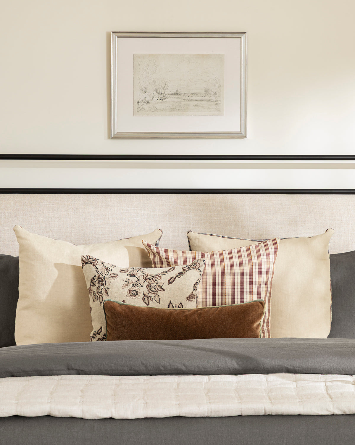 A neatly made bed with gray and cream bedding, beige plaid and floral pillows, sits beneath the French Countryside Sketch in a silver-finished frame on a light wall.