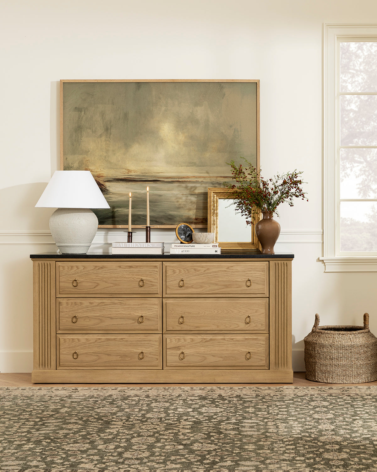 A wooden dresser with eight drawers stands against a cream wall. On top are a lamp, candles, books, a clock, and a framed Misty Blue Headland giclee print. A vase of flowers, gold-framed mirror, and basket complete the scene, by McGee & Co.