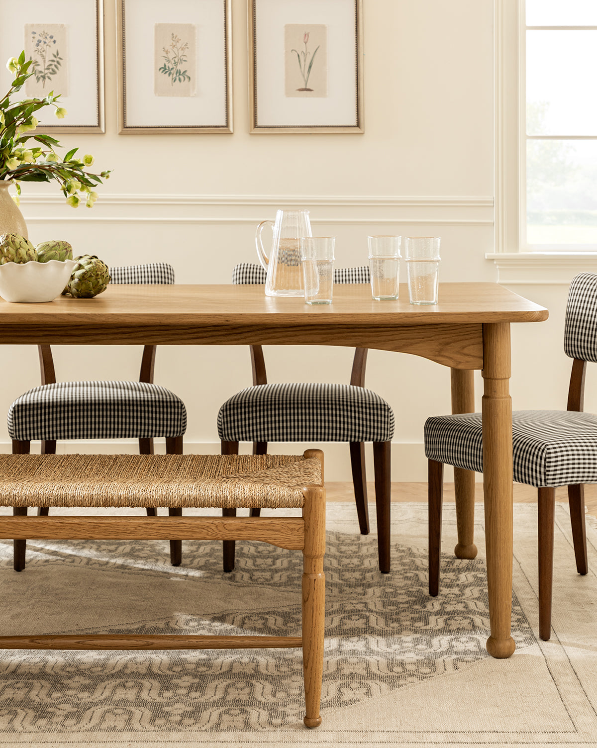The Phipps Dining Table is paired with a pitcher, four glasses, and a bowl of artichokes, surrounded by plaid-upholstered chairs and a woven bench in a bright room featuring framed botanical art and a window by McGee & Co.