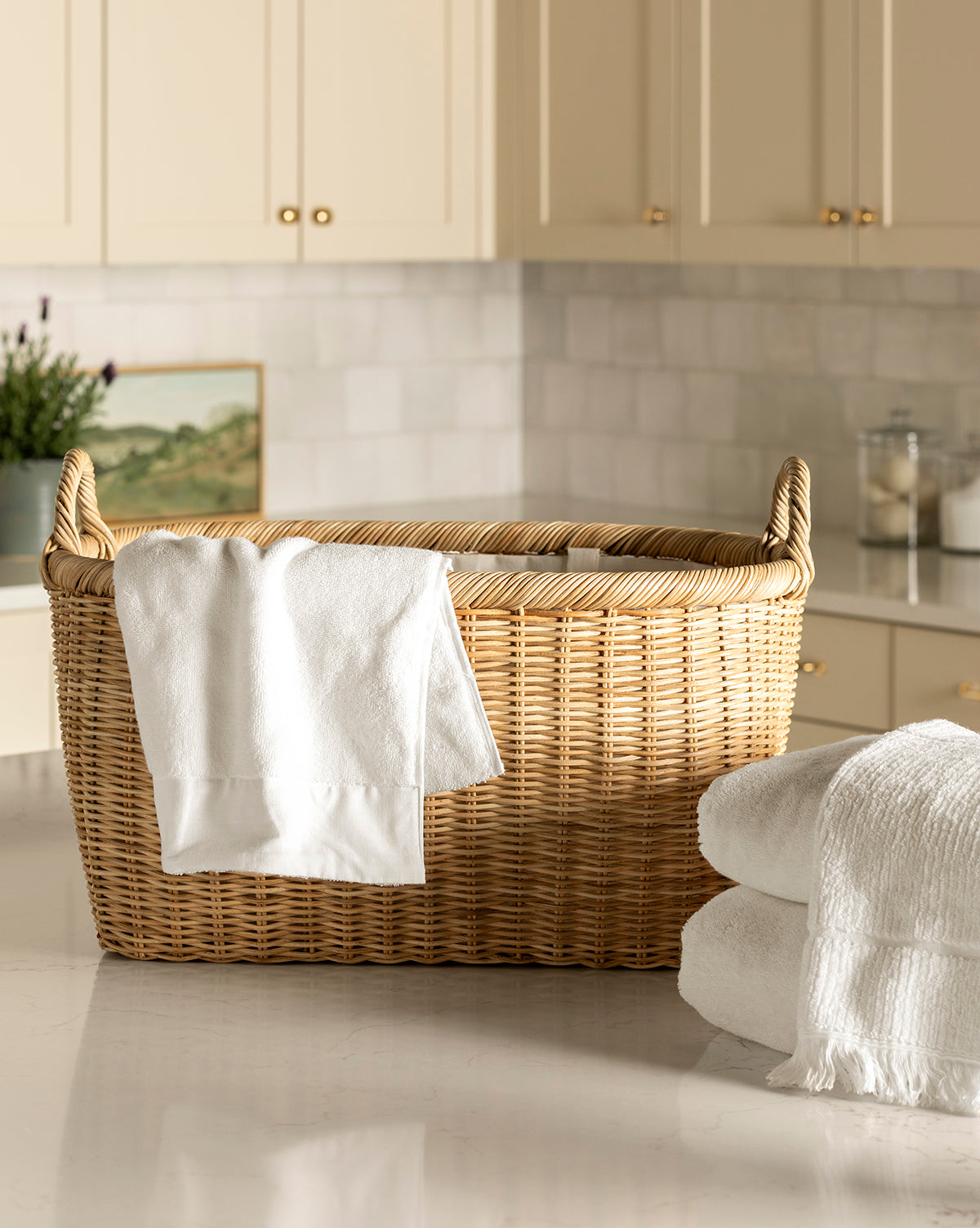 The Fairway Laundry Basket with a white towel draped over its side sits on a kitchen counter next to neatly folded white towels, with cream cabinets and jars in the background, by McGee & Co.