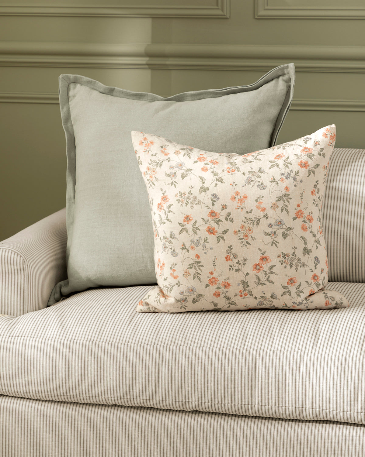 Two Liam Double Flange Pillow Covers—a solid light gray and a cream with orange floral print—decorate a beige striped sofa in front of a pale green paneled wall, by McGee & Co.