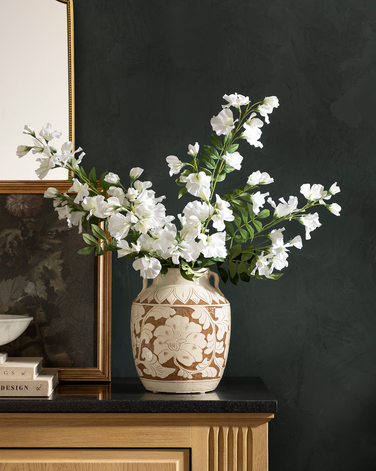 The Blaise Vase, featuring an ornate floral pattern, holds white flowers and green leaves on a black countertop beside books and a framed mirror, adding timeless charm to your home decor against a dark textured wall, by McGee & Co.