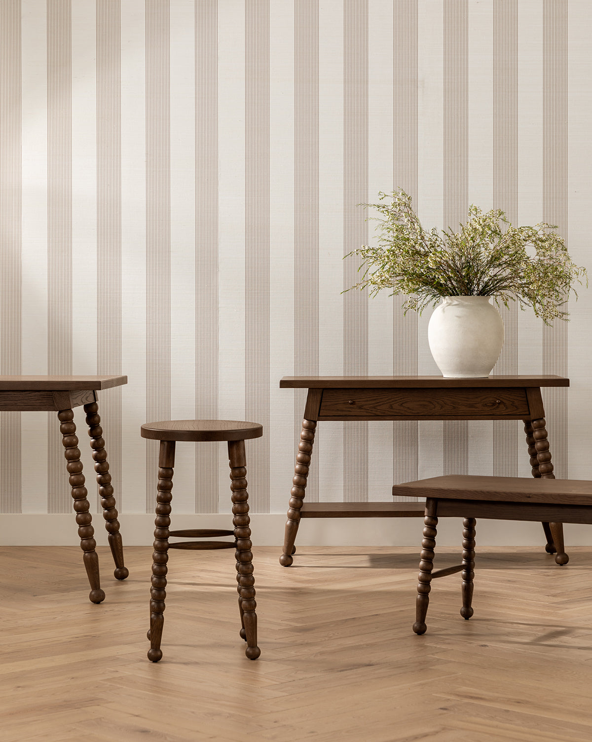 A minimalist room with light striped wallpaper, wooden furniture including the Heathcliff Bench with spindle legs, and a white vase with green foliage on a wooden table. The warm herringbone floor completes the look, by McGee & Co.