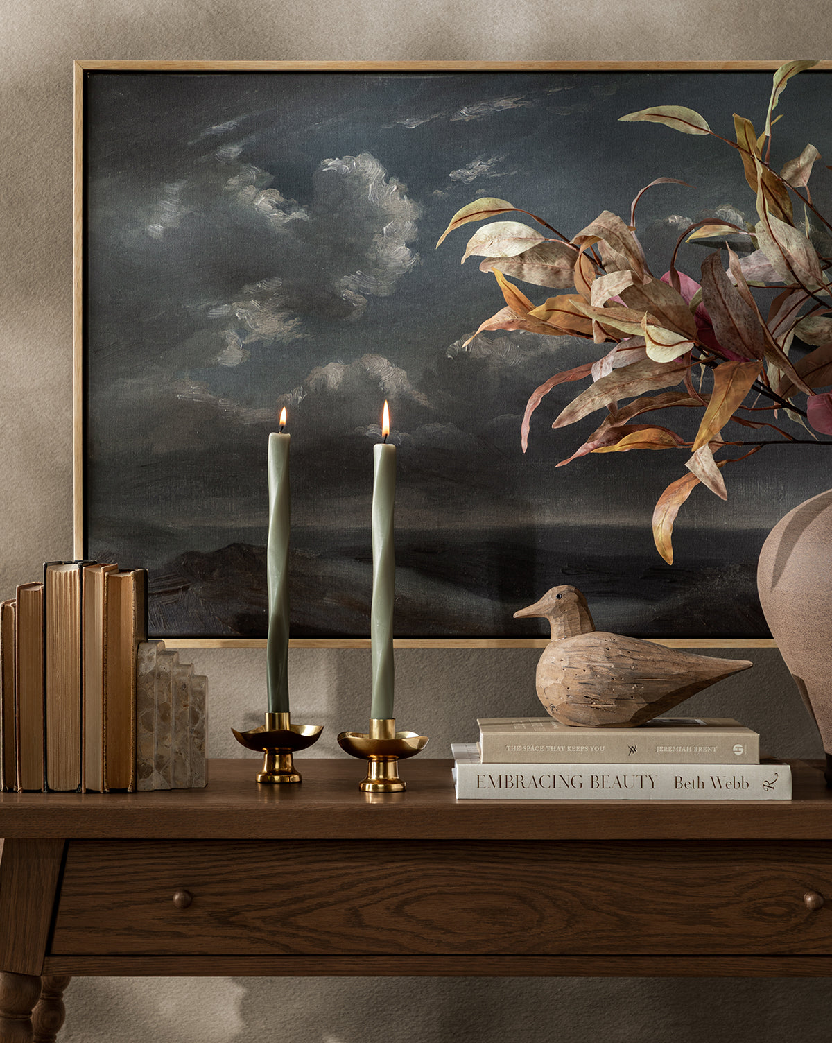 A wooden table displays stacked books, two lit green candles in Cosima Taper Holders, a wooden bird sculpture, and a vase with leafy branches. A moody landscape painting on the wall complements the elegant decor, by McGee & Co.